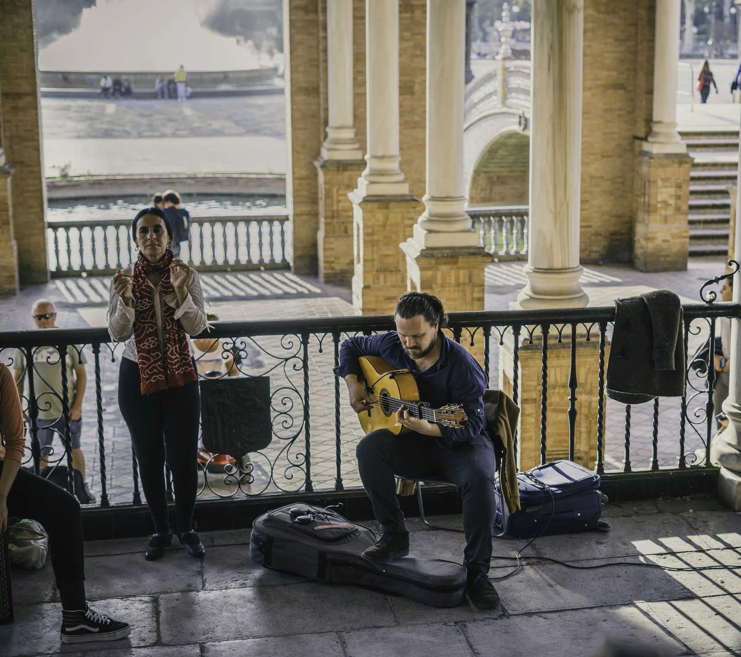 Musician playing guitar in public setting with woman clapping, arched architecture background.