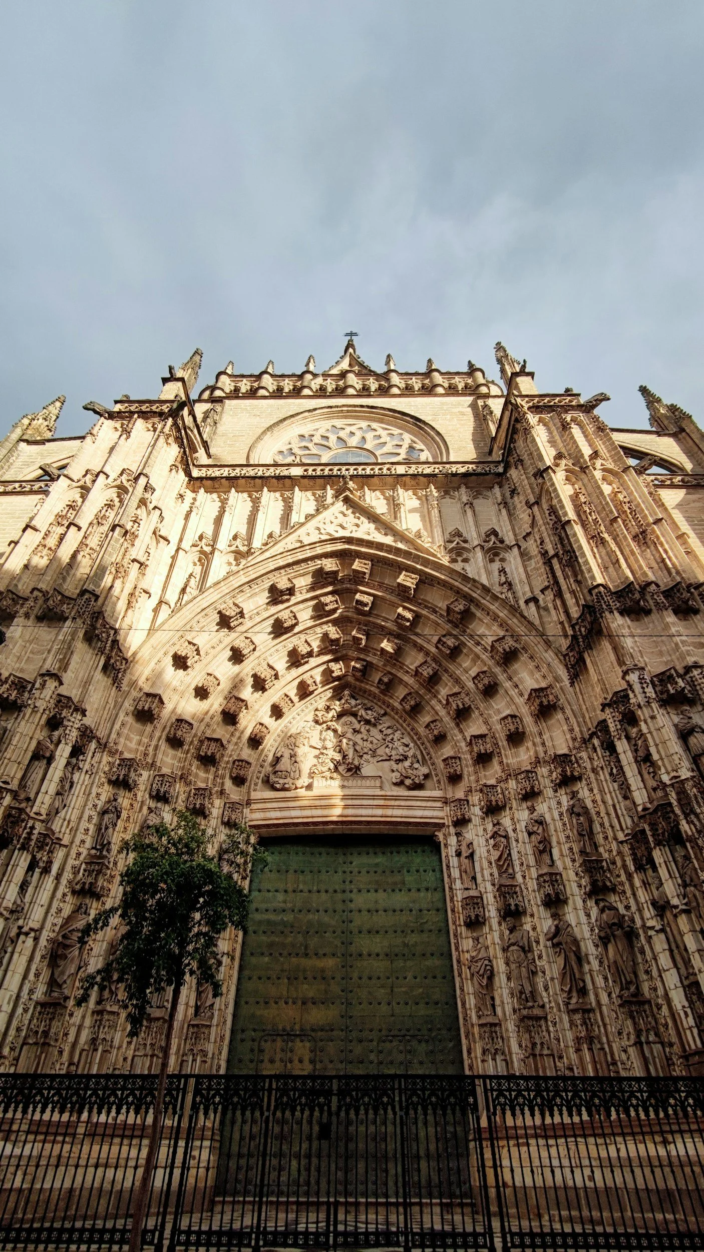 Gothic cathedral facade with ornate carvings and large green door, under a cloudy sky.