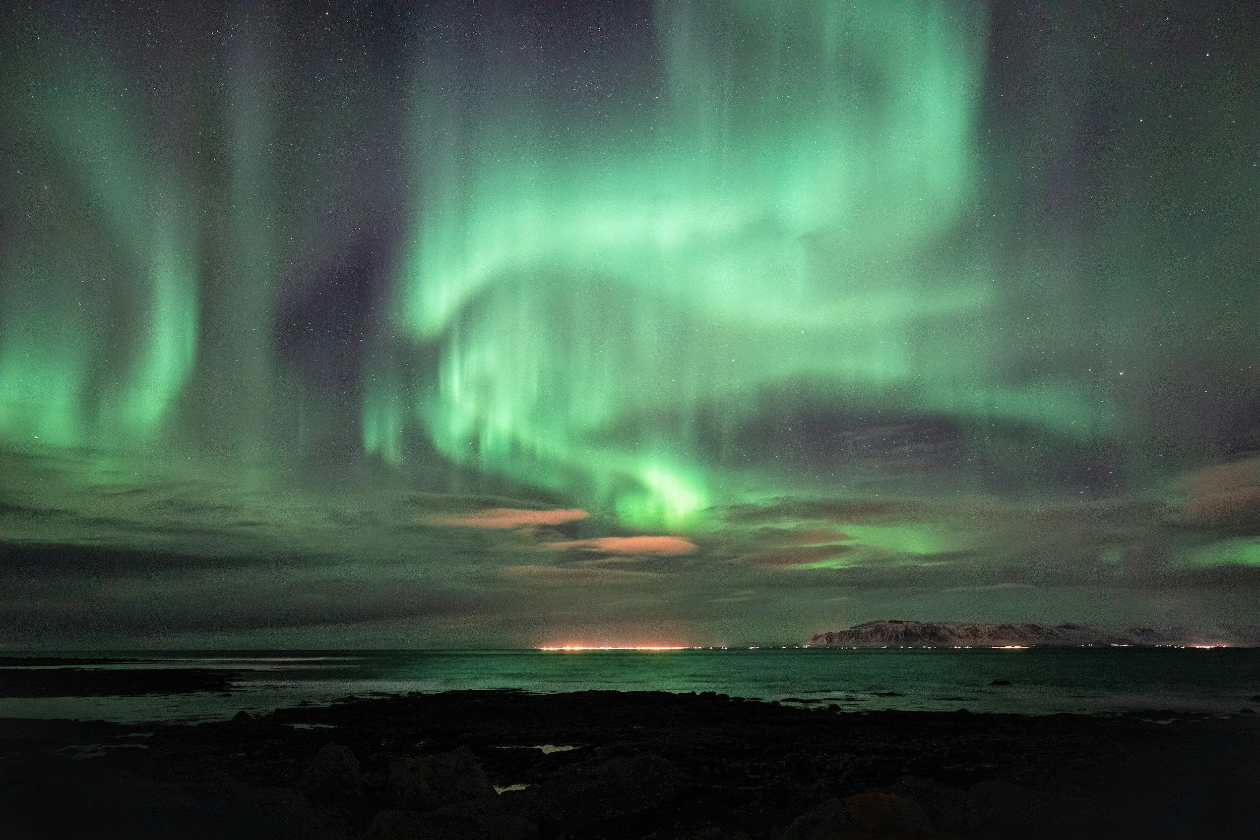 Northern Lights over a coastal landscape with water and distant mountains.