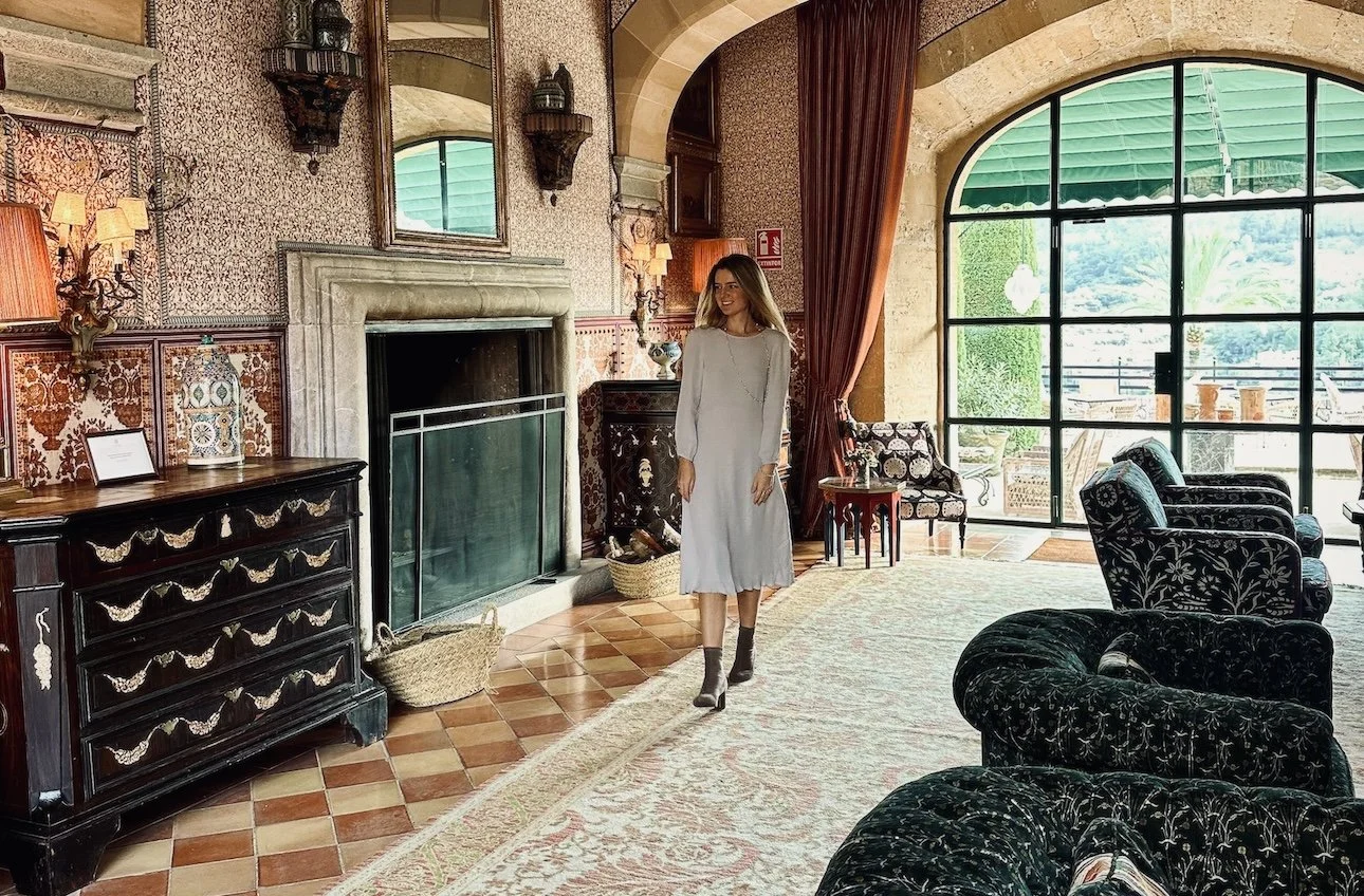 A woman in a light gray dress with black socks and heels walking inside a cozy vintage-style room with patterned walls, a fireplace, and large glass doors leading to a balcony with green scenery.