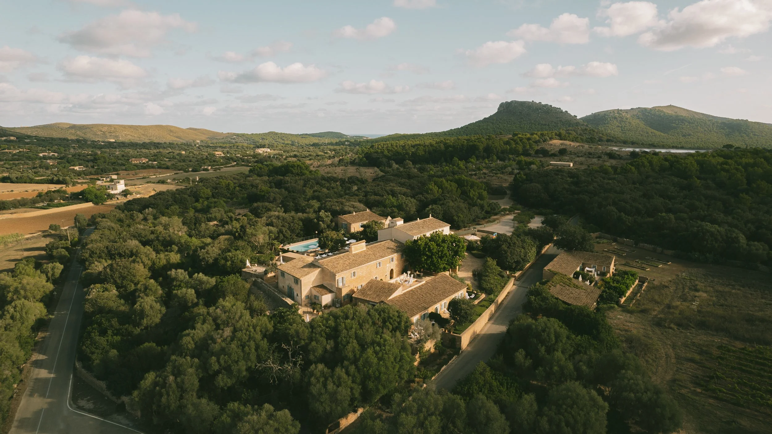 Aerial view of a countryside estate with several buildings, a swimming pool, surrounded by trees, with hills in the background.