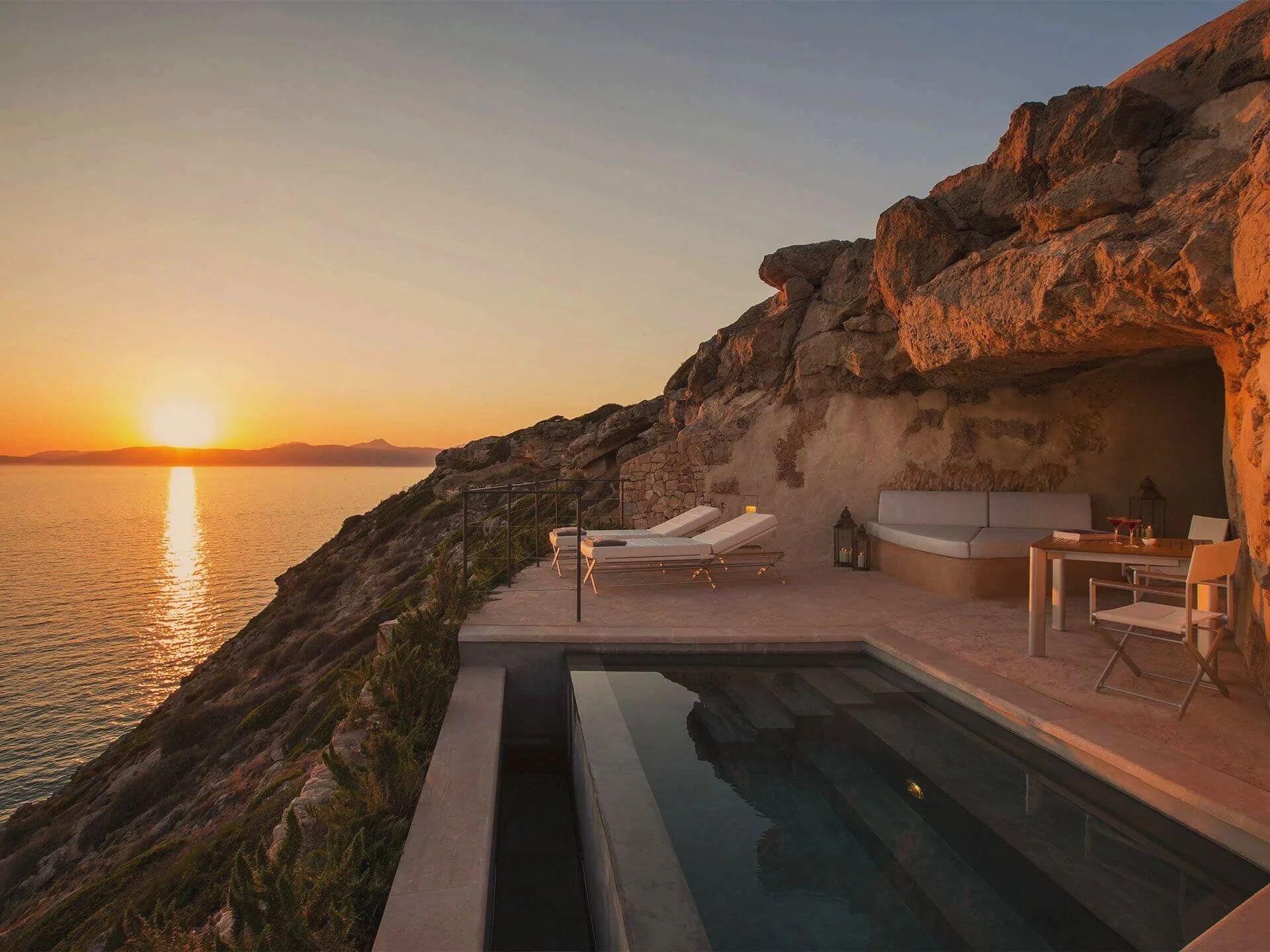Luxury cliffside patio with pool, table, and lounge chairs overlooking ocean at sunset.