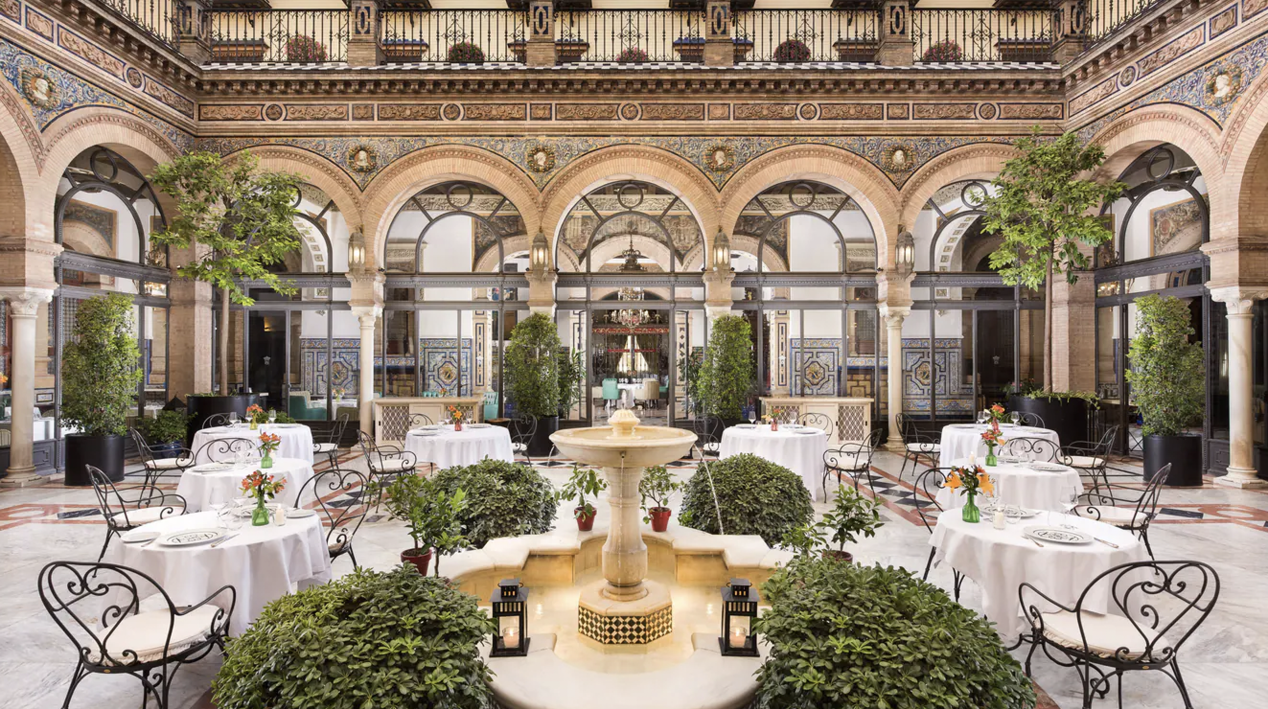 Elegant outdoor restaurant courtyard with white tablecloth-covered tables and wrought iron chairs, surrounded by potted plants and trees, featuring a central fountain and decorative archways.