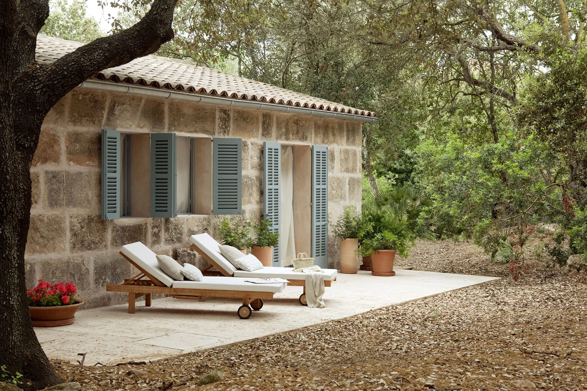 A rustic stone house with blue shutters and a tiled roof, surrounded by trees and potted plants, with two white lounge chairs on a patio.
