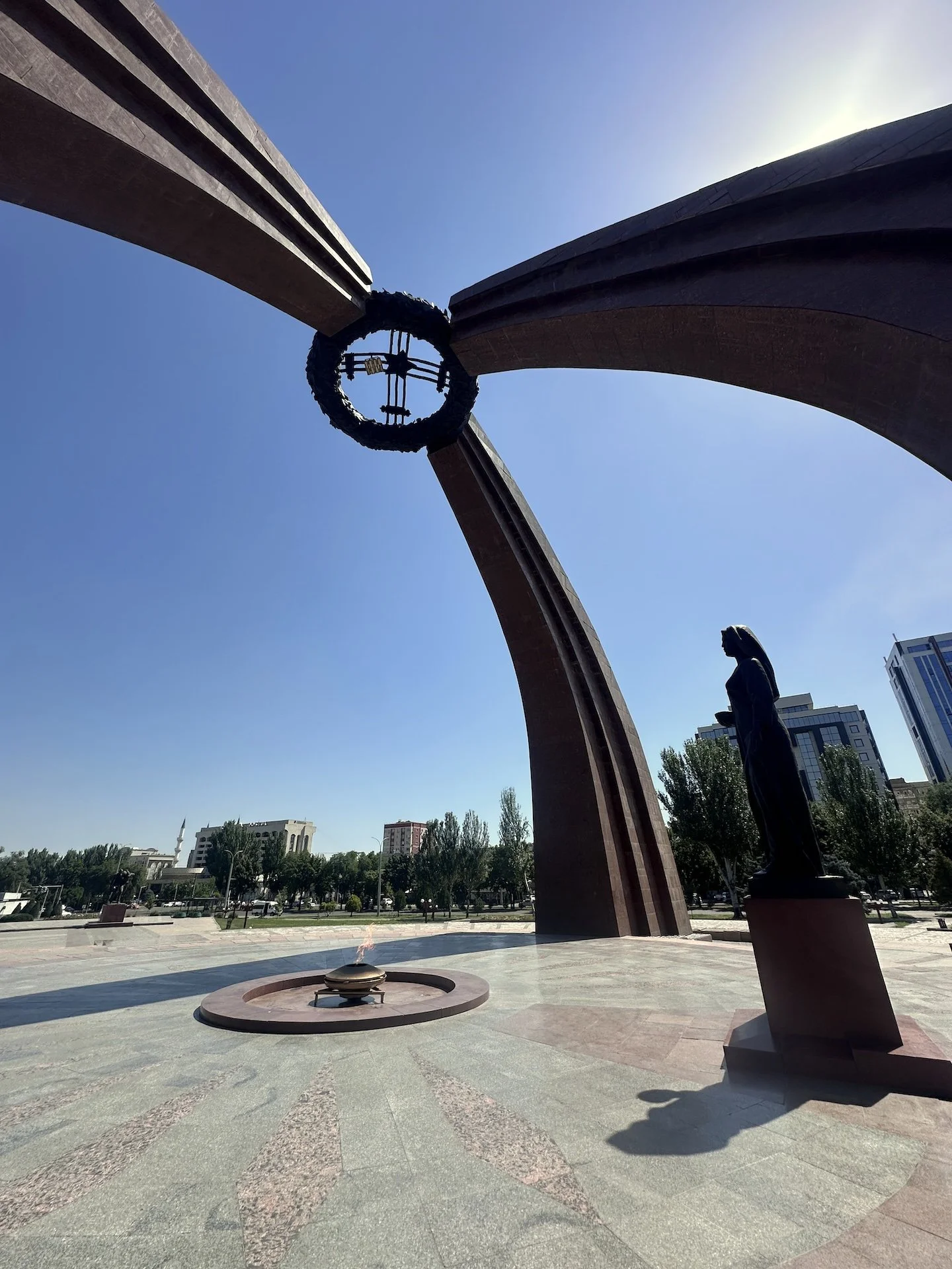 A monument with tall curved structures, a statue of a woman, and an eternal flame at the center, set in a park with trees and buildings in the background.