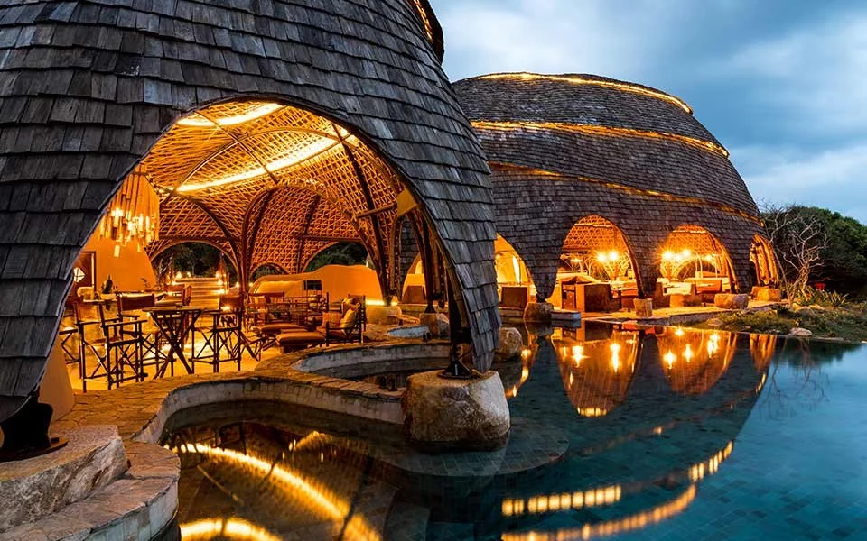 Luxurious safari lodge with thatched roof, warm interior lighting, and reflection in a pool at dusk.