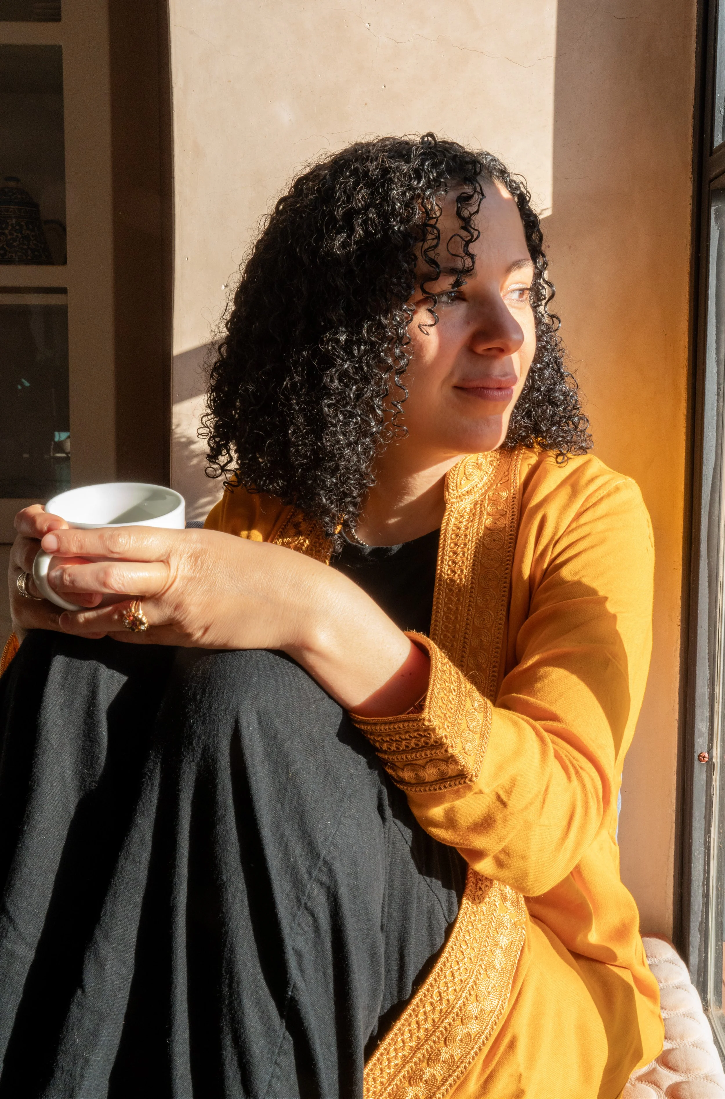 Woman sitting by a window, holding a white cup and wearing an orange embroidered jacket with curly hair.