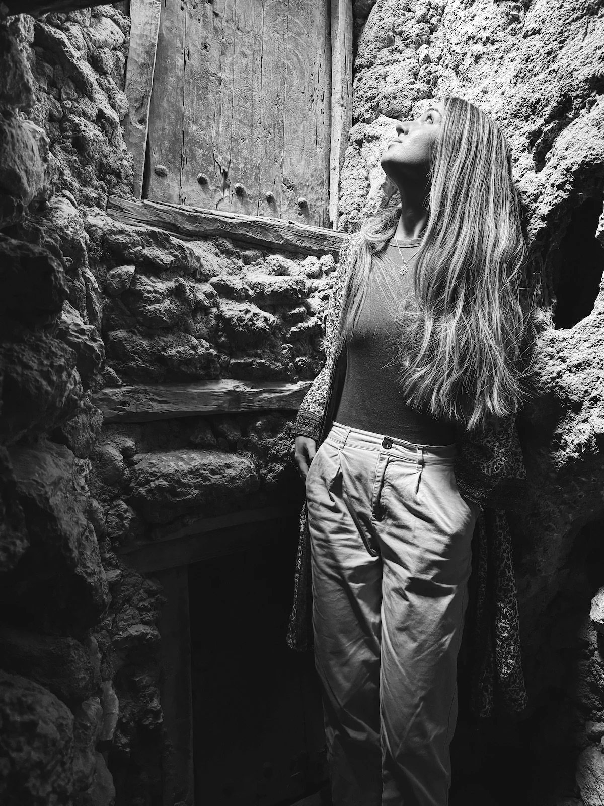 A woman with long, wavy hair standing in a small, stone-walled space, looking up, wearing a short-sleeved top and high-waisted pants.