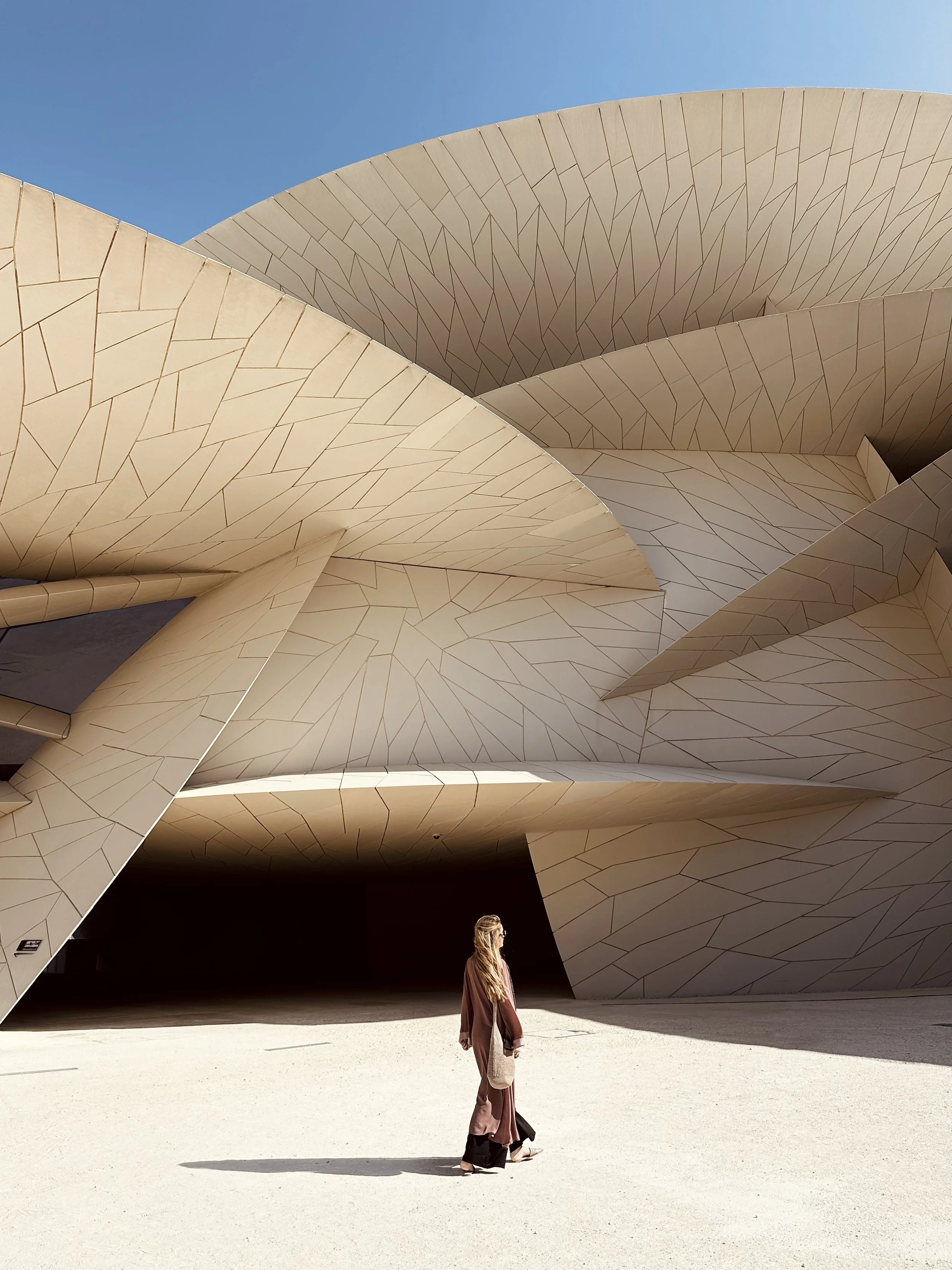 A woman walking in front of a modern, abstract building with beige, angular, overlapping panels under a clear blue sky.
