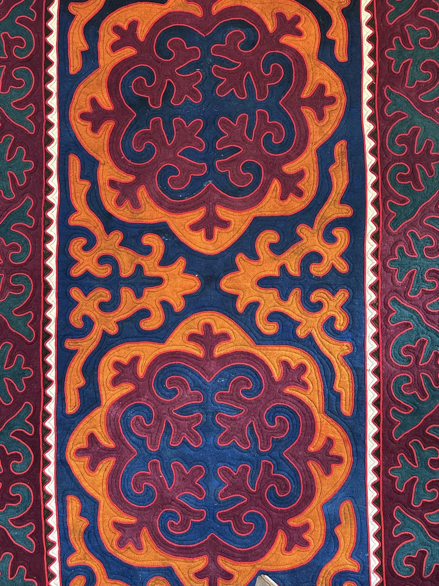 A colorful textile with intricate patterns, including a central symmetrical design with swirling shapes in orange, pink, and dark blue, bordered by narrower bands with repeated motifs in red, green, and white.
