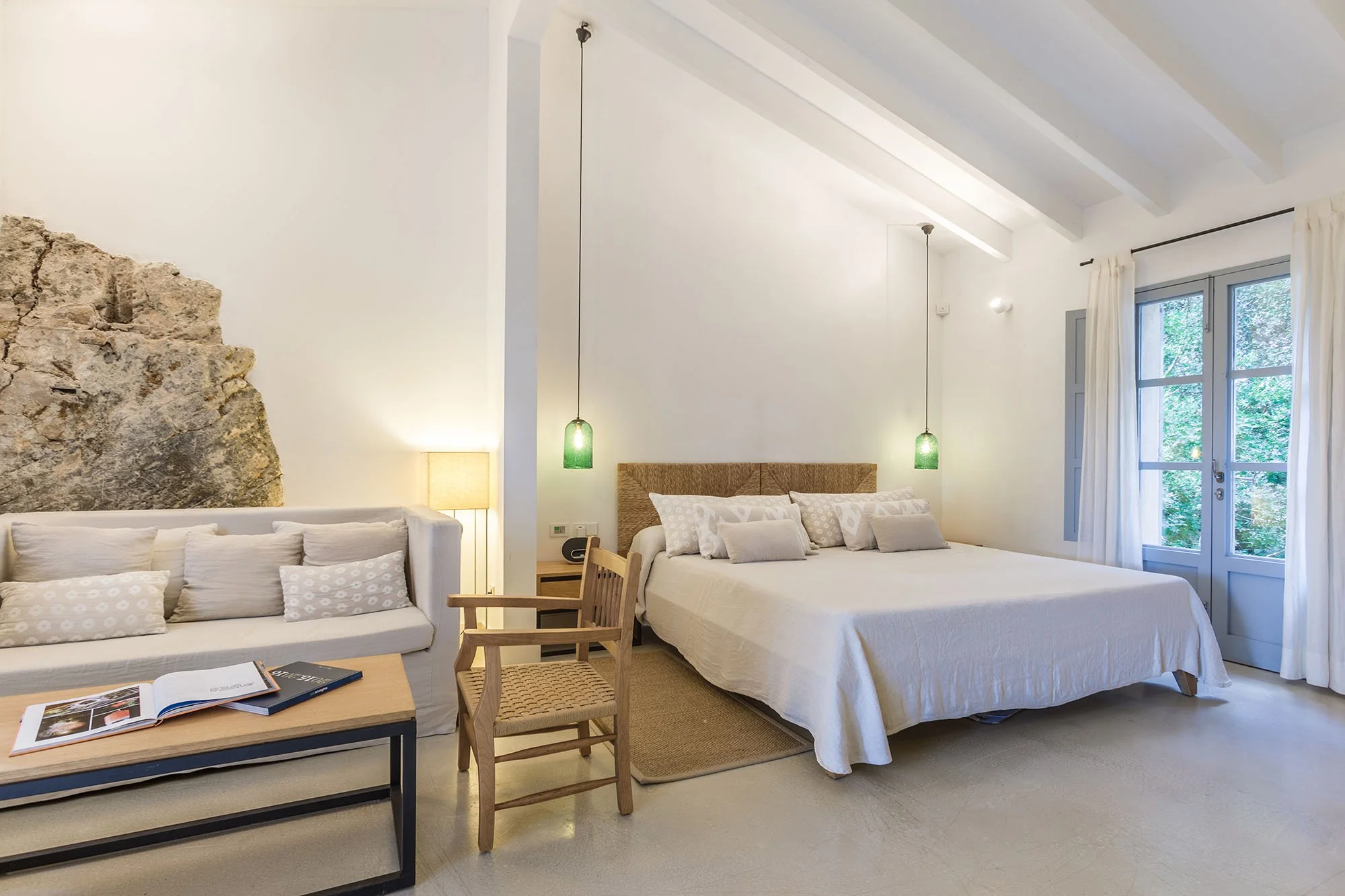 A bright bedroom with white walls, featuring a large bed with white bedding and multiple pillows, a built-in stone wall detail, a wooden chair, a sofa with pillows, a wooden coffee table with books, a nightstand with a lamp, and glass doors with white curtains leading outside to greenery.