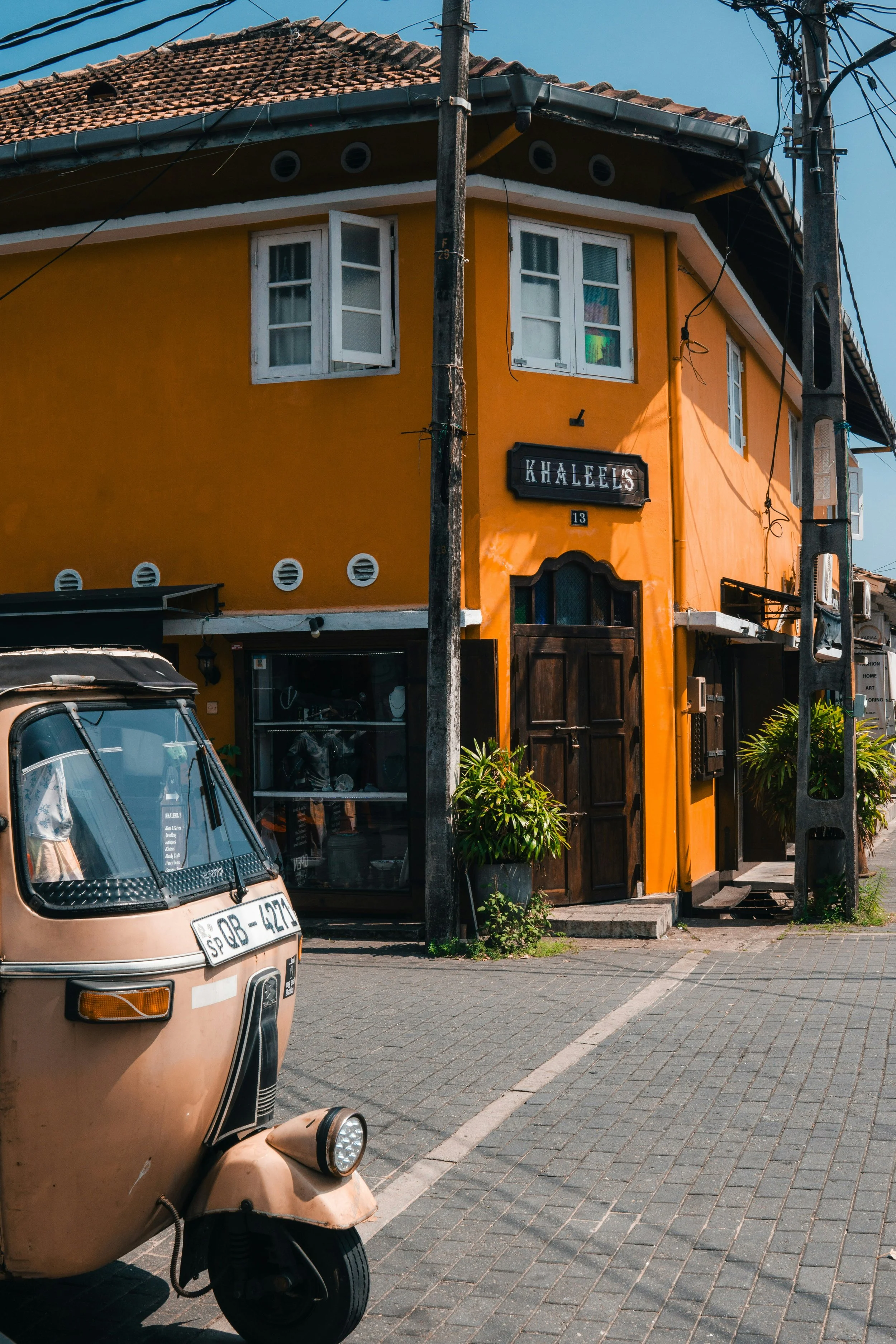 Bright yellow building with a storefront named Khaleel's, located on a corner with a cobblestone street. A tuk-tuk is parked in front of the building.