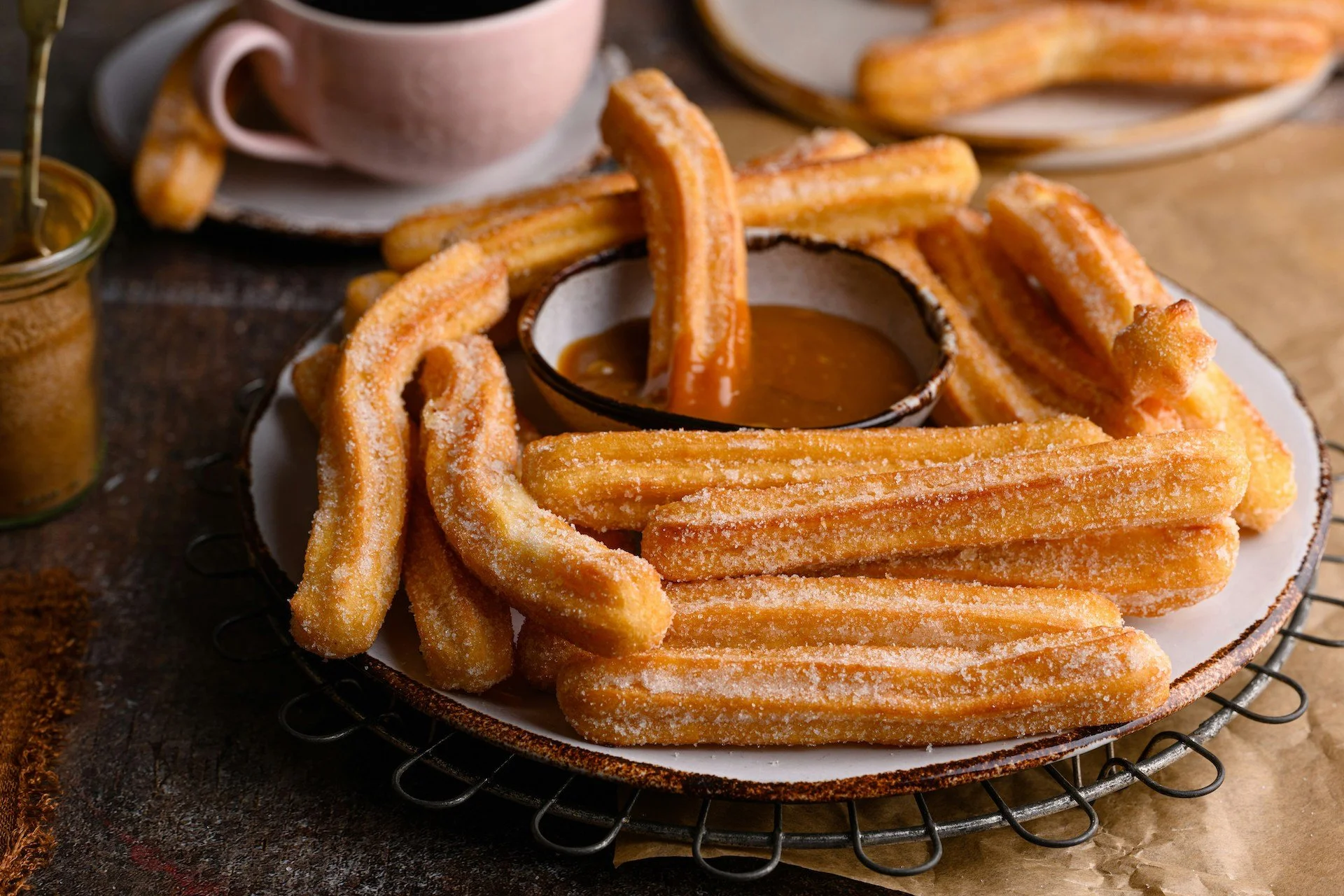 Plate of sugar-coated churros with caramel dipping sauce, a cup of coffee in the background.