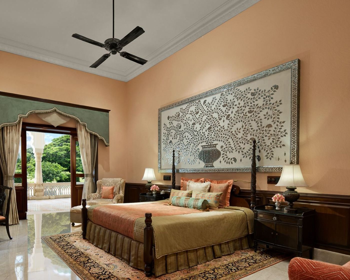 Luxurious bedroom with a large bed, ornate wall art, and elegant furniture, featuring a ceiling fan, lamps, and an open view to a balcony with greenery.