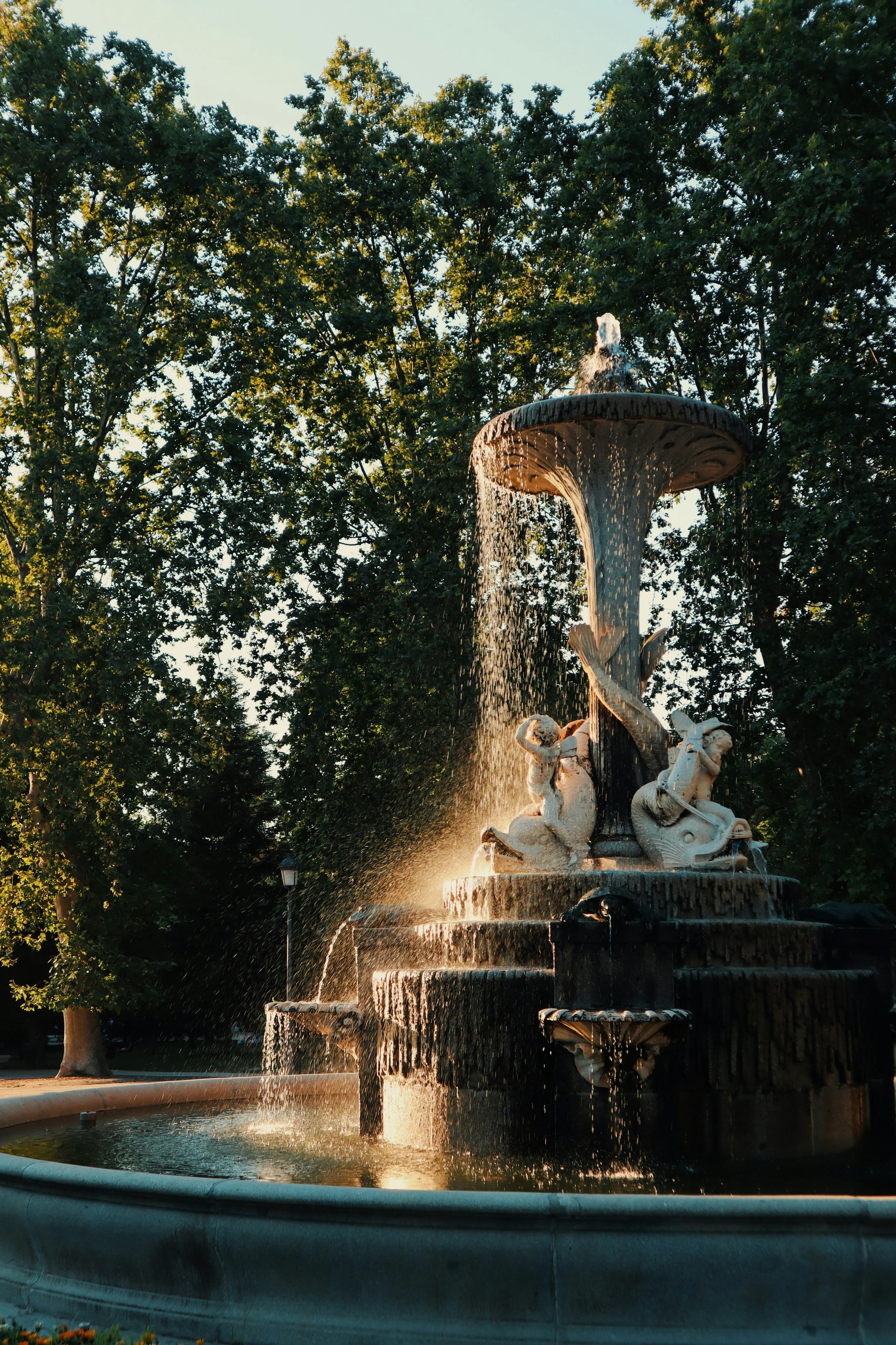 Ornate stone fountain with water cascading over sculptures framed by trees