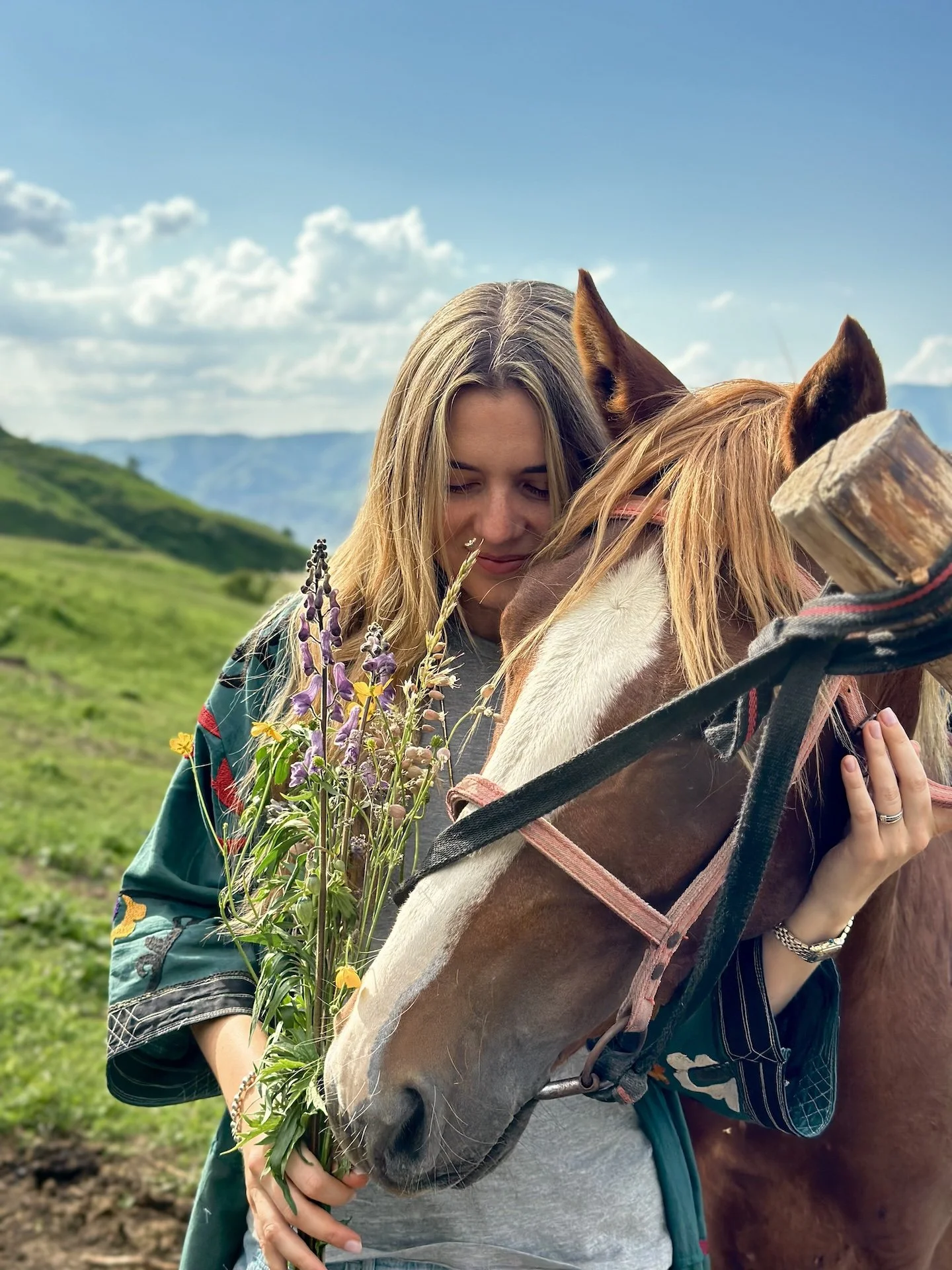 A woman with blonde hair hugging a brown and white horse, holding a bouquet of wildflowers, outdoors with mountains and a partly cloudy sky in the background.