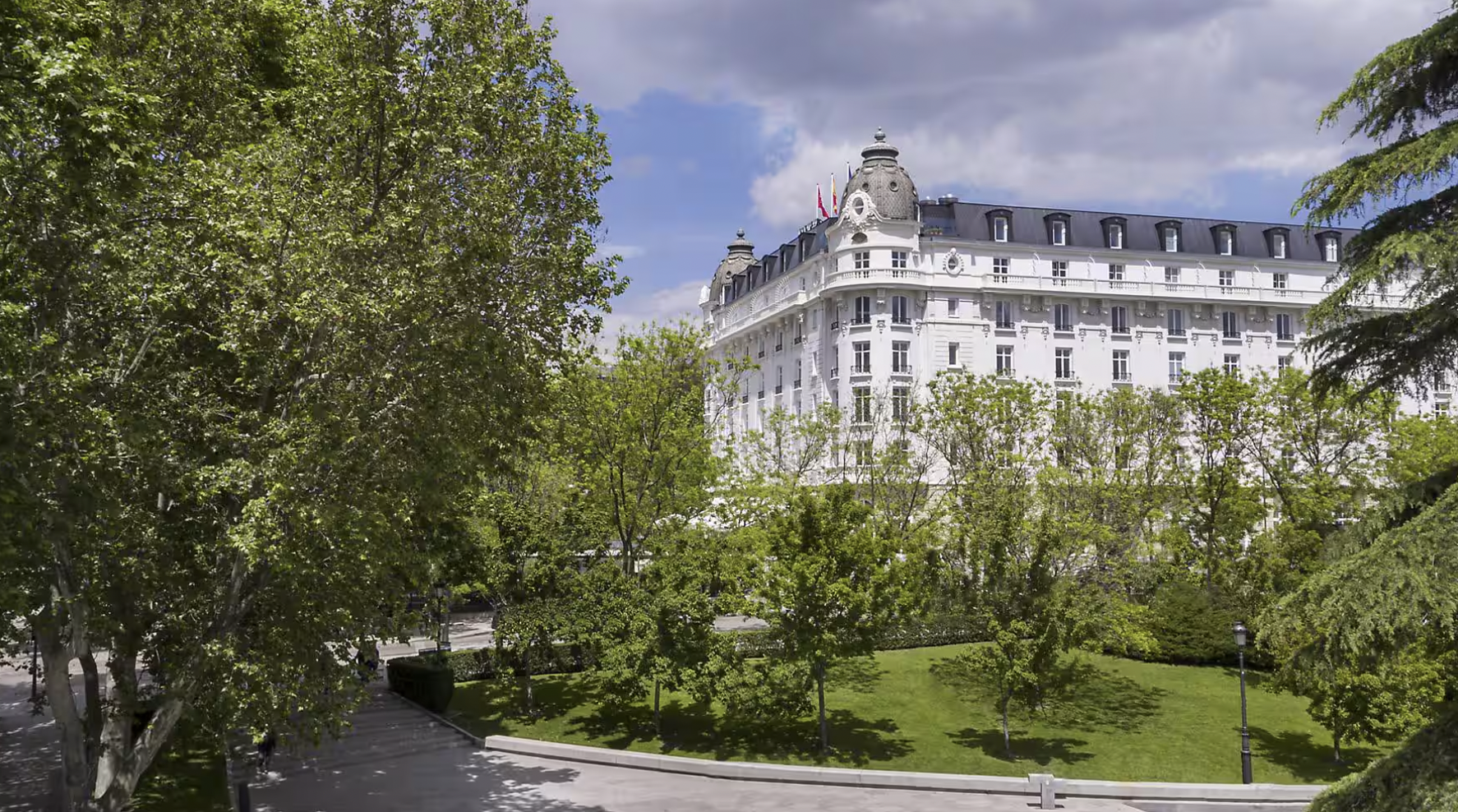 Historic white building with decorative architecture behind lush green trees under a partly cloudy sky.
