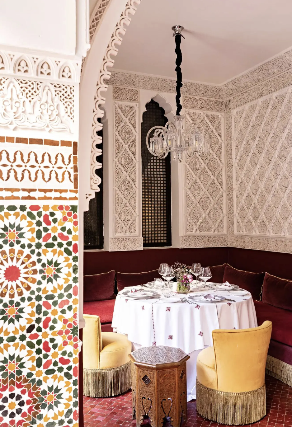 Elegant dining room with a round table, set for a meal with wine glasses, plates, and flowers, surrounded by plush chairs and intricate Moroccan-style wall decor.