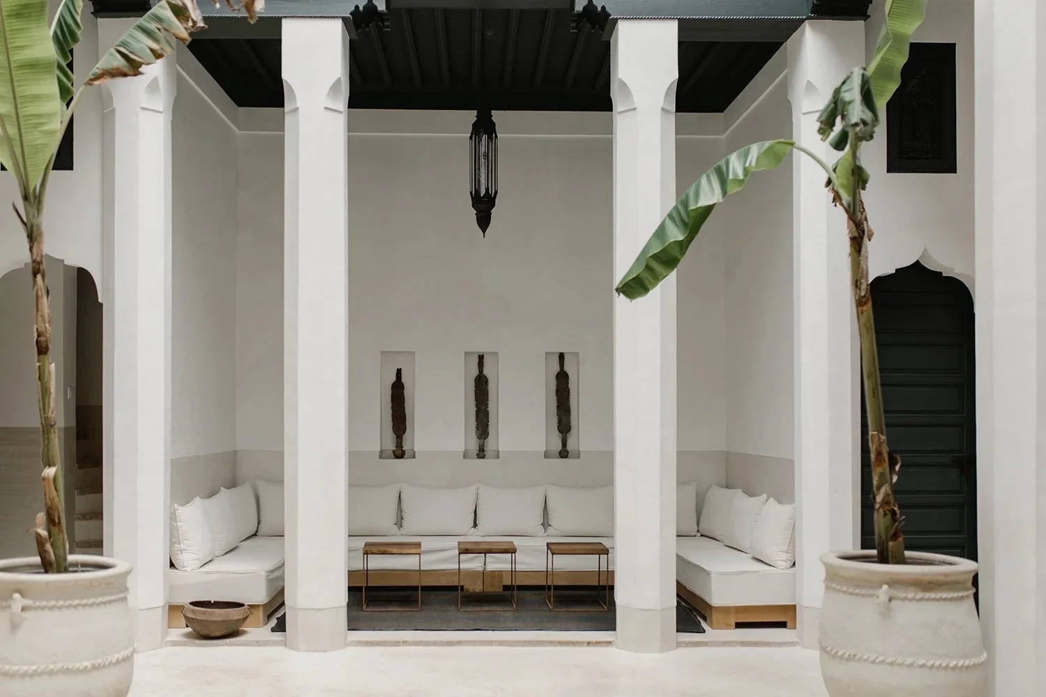 Luxurious courtyard with tropical plants, a reflecting pool, wicker lounge chairs with orange cushions, lanterns, and a seating area with white curtains under a warm evening sky.
