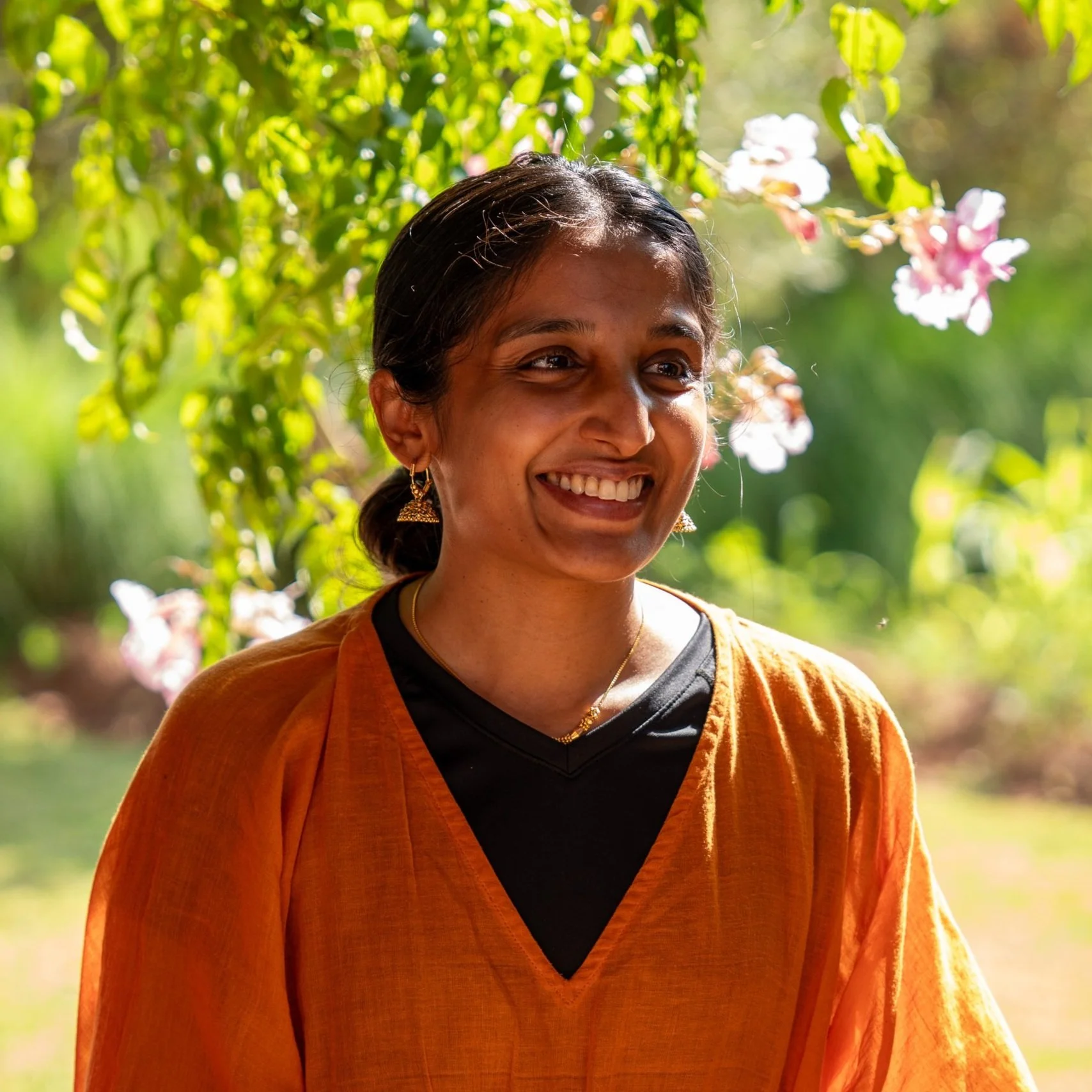 Smiling person outdoors wearing an orange outfit and gold earrings, with green foliage and pink flowers in the background.