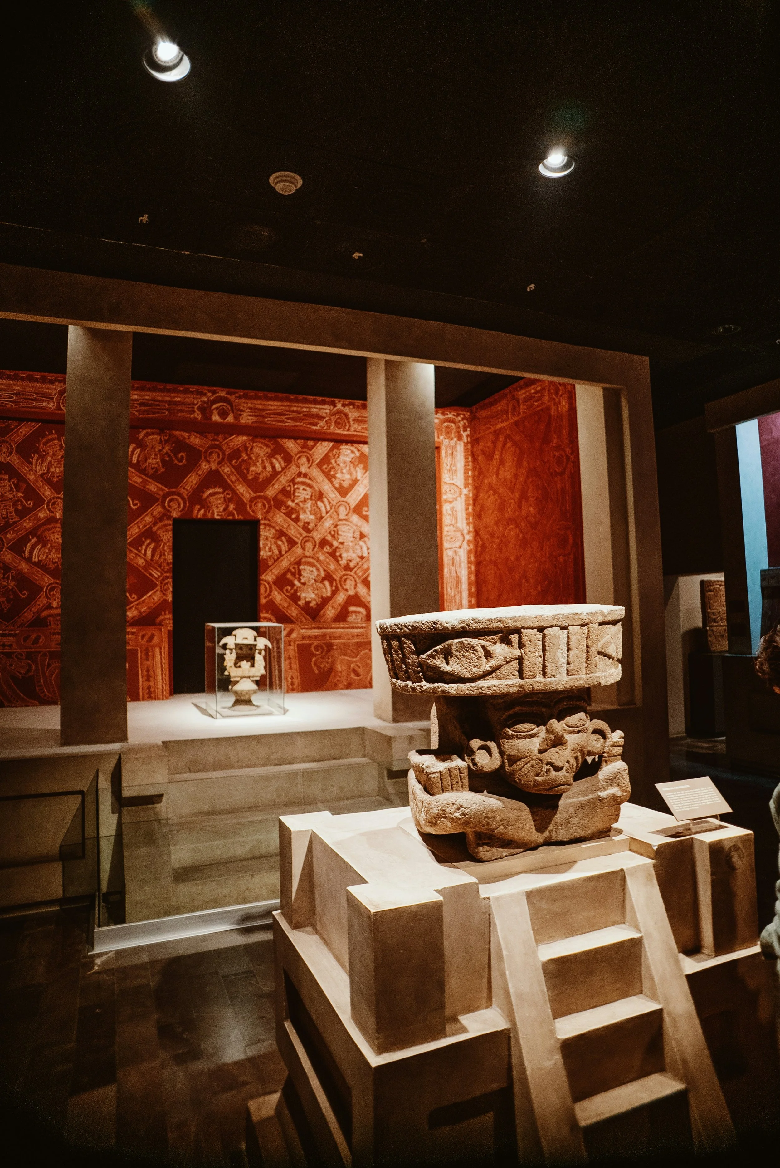 Ancient stone sculptures displayed in a museum exhibit with intricate red wall patterns and subdued lighting.
