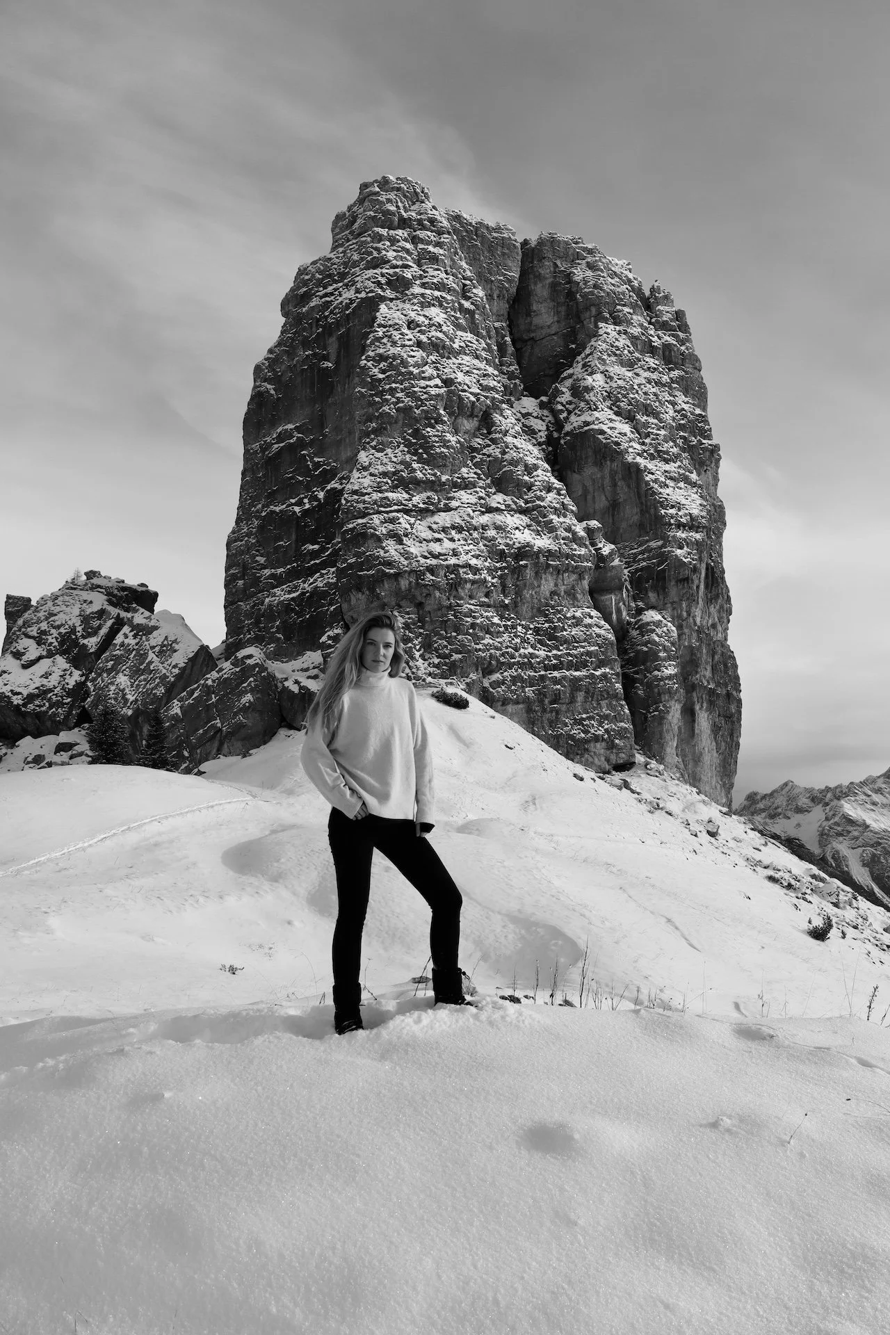 A woman standing on snow in front of a large, snow-covered rocky mountain in a black and white photo.