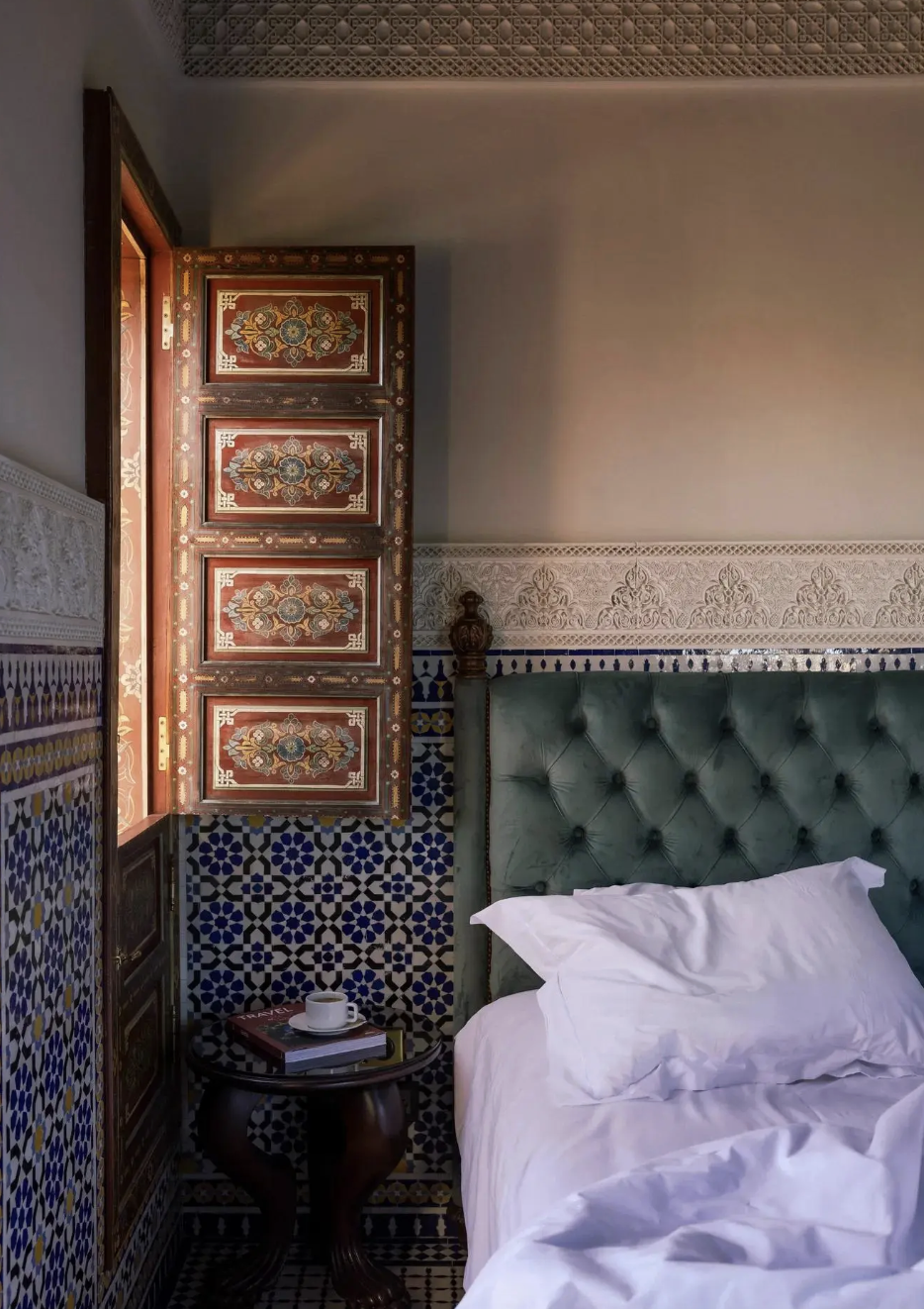 A cozy bedroom corner with a green tufted headboard, a white bed with an unmade pillow, a small dark wooden side table with a cup on top, and a decorative wall with intricate Moroccan-style tile work and a carved wooden window shutter.