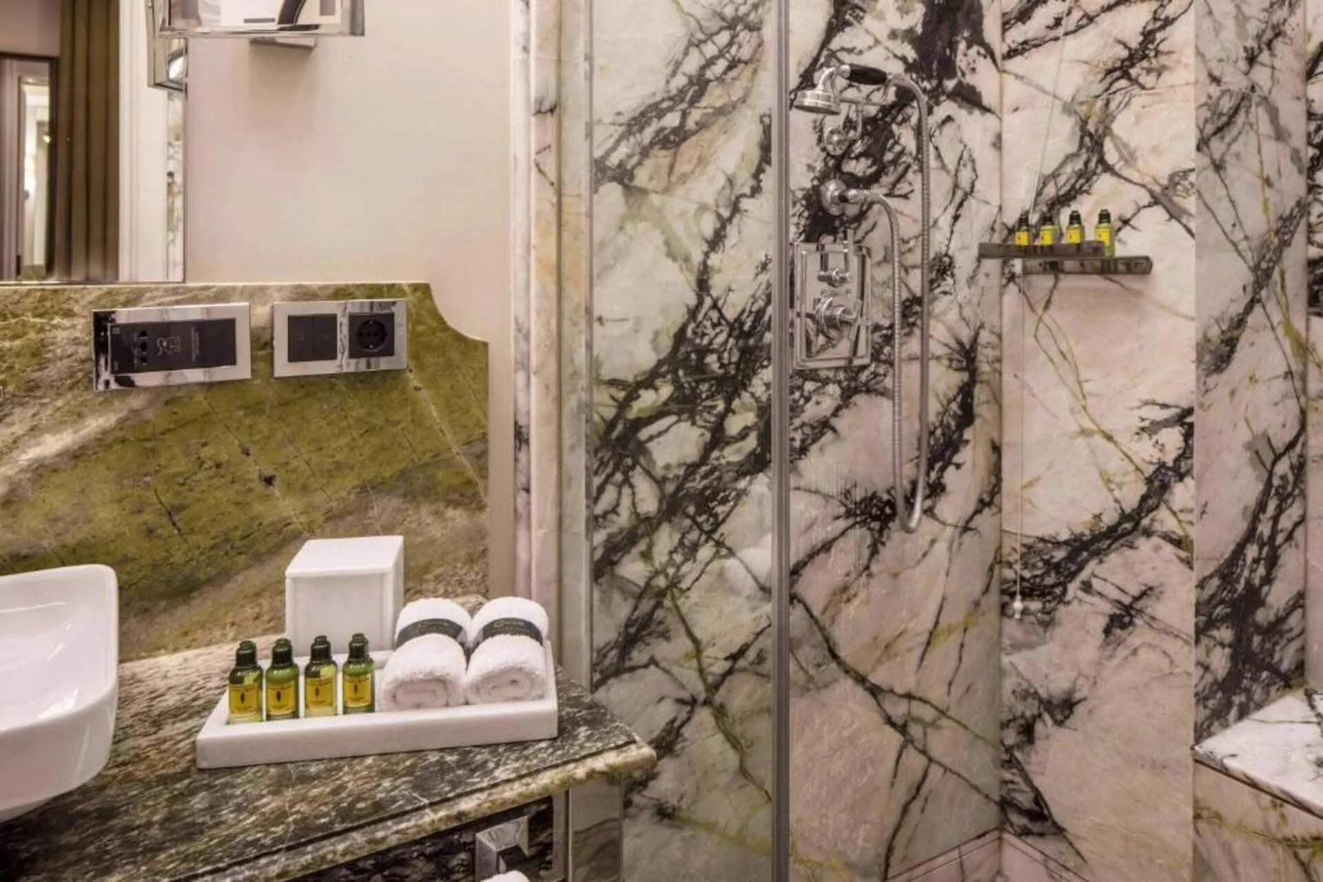 Luxurious bathroom with marble walls, a walk-in shower with a head and controls, a shelf with toiletries, and a sink area with towels and toiletries.