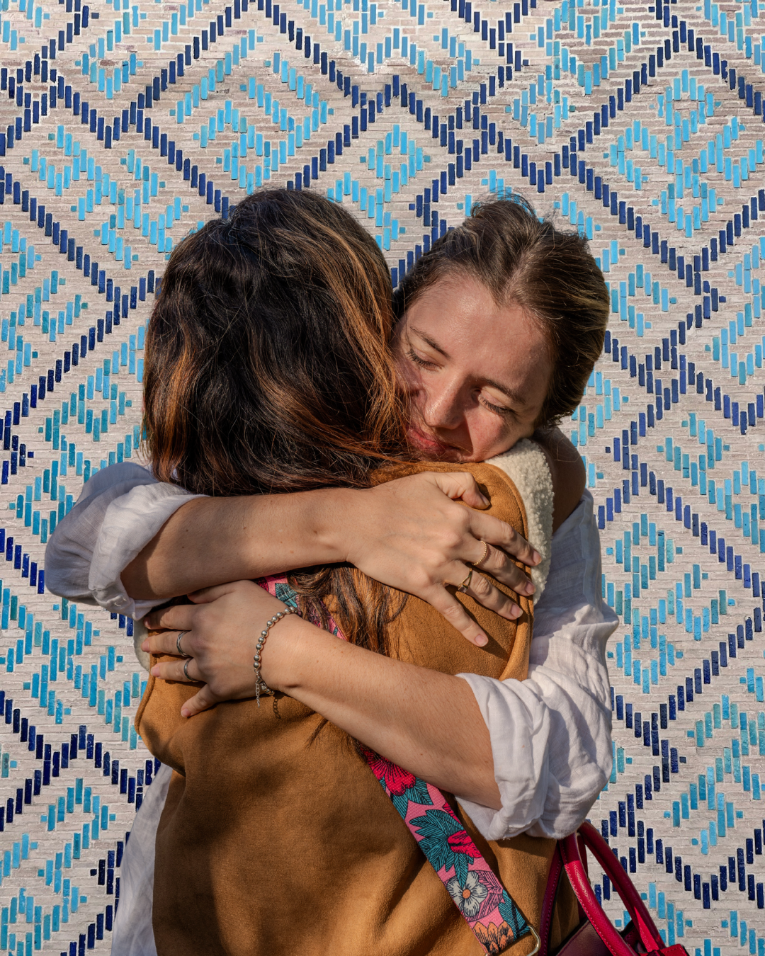 Two women hugging warmly against a patterned blue and white background, one with curly hair and the other with straight hair and a floral strap.