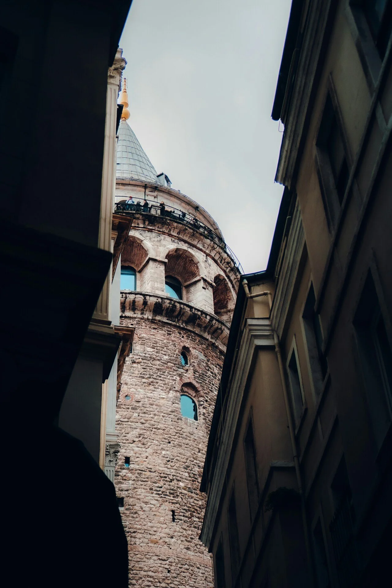 View of a tall, round stone tower with arched windows, seen from a narrow alley between two buildings under a cloudy sky.
