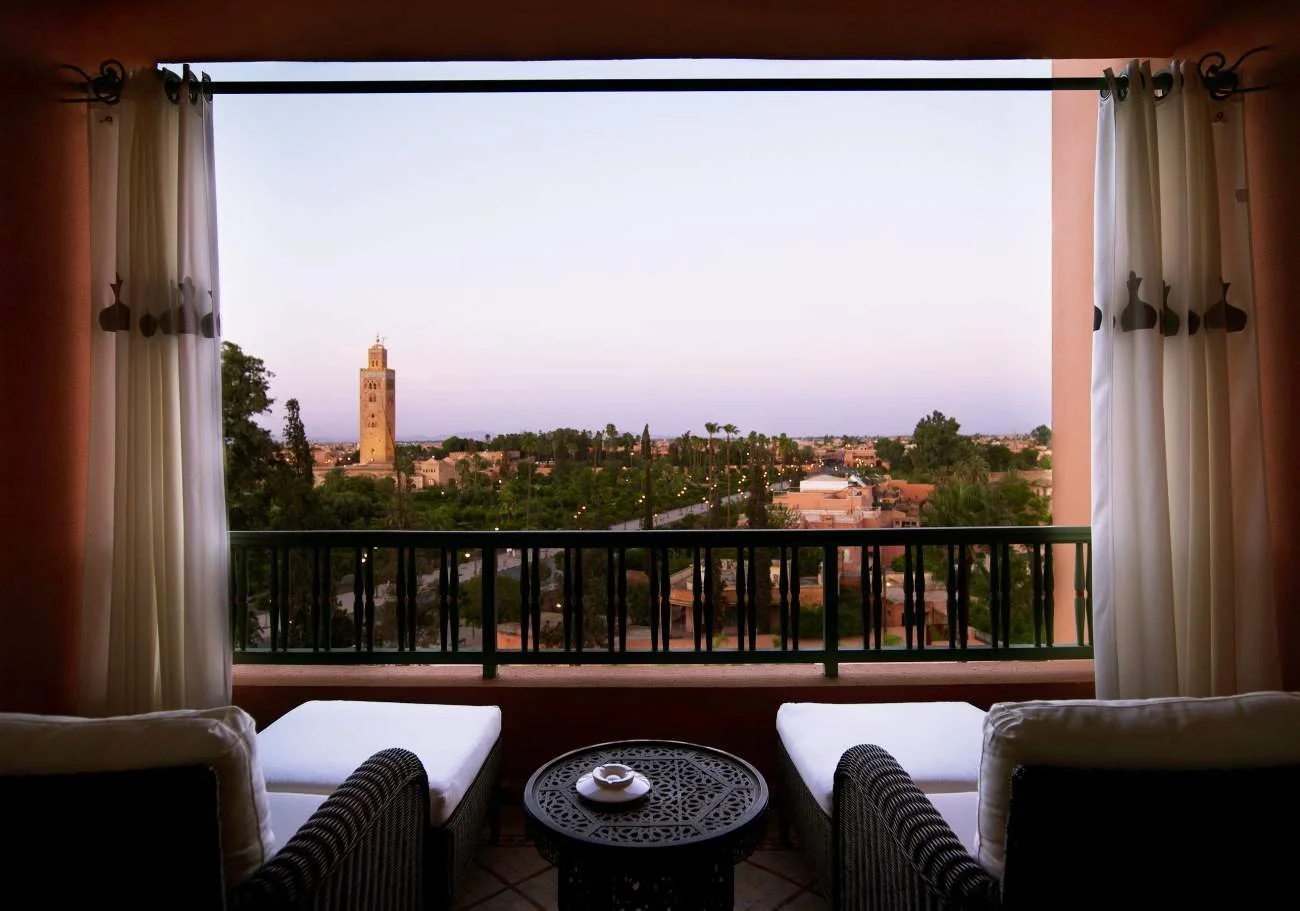 Balcony view with wicker chairs and table overlooking a cityscape with a tall tower and greenery at sunset.