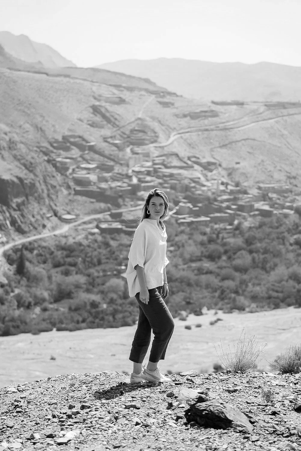 A woman standing outdoors on rocky ground with mountainous terrain and a small town in the background.