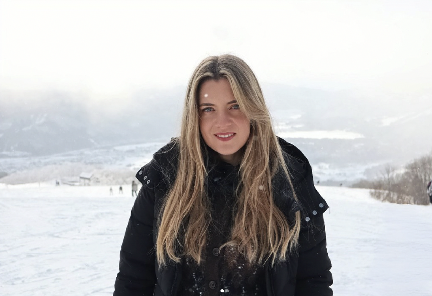 A young woman with long blonde hair and blue eyes standing outside in a snowy landscape during daytime, wearing a black jacket with snow on her shoulders.