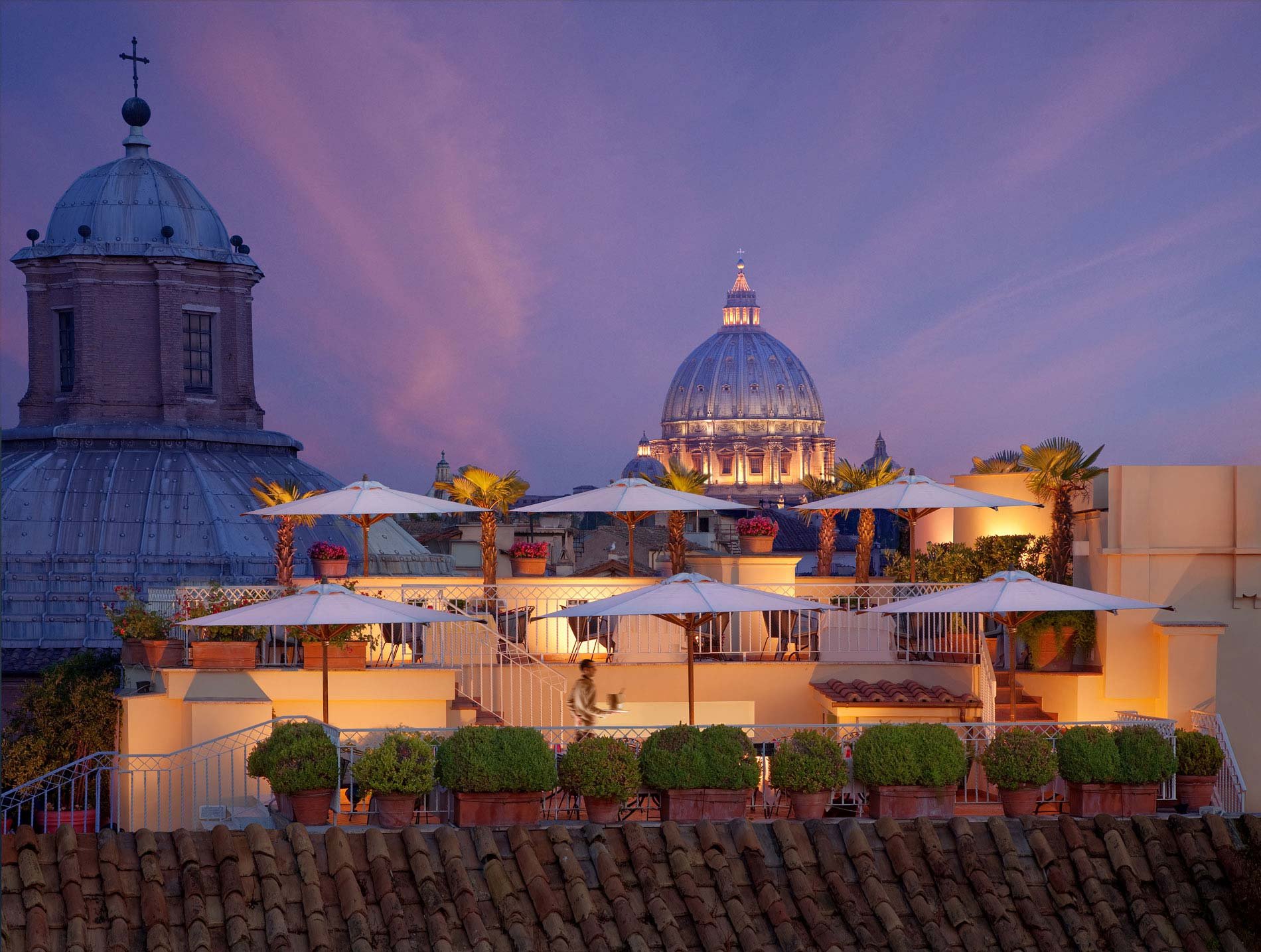 Rooftop terrace with umbrellas and potted plants, view of St. Peter's Basilica, evening sky