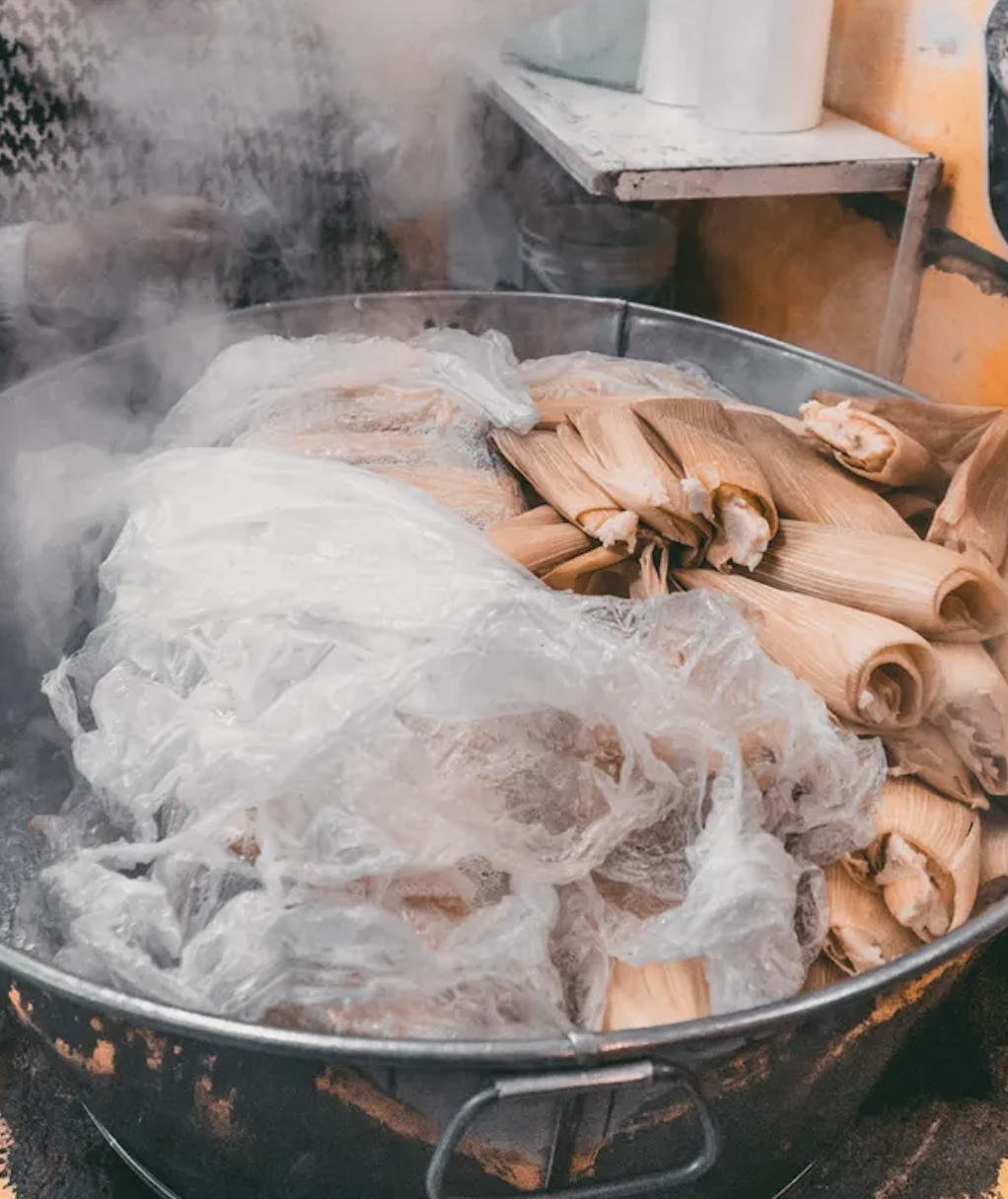 Steaming tamales wrapped in corn husks inside a pot, partially covered with a plastic sheet, emitting visible steam.