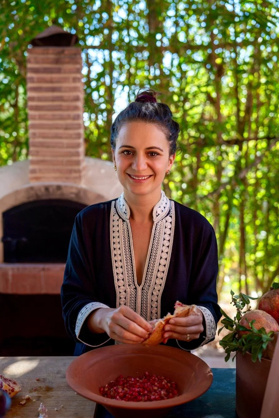 A woman in a traditional embroidered top preparing pomegranates outdoors by a brick oven, with a bowl of pomegranate seeds on the table.