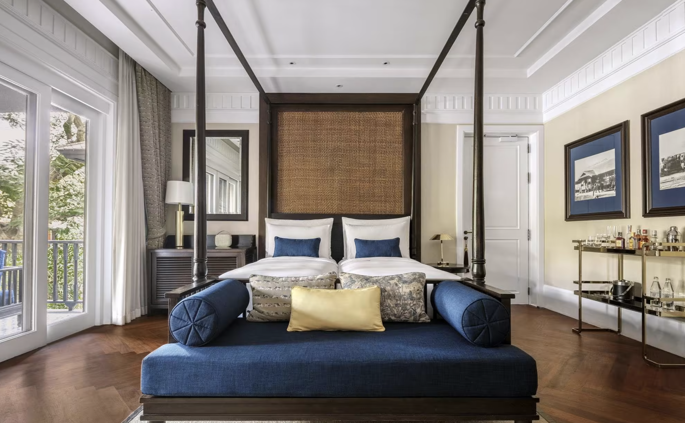 Elegant hotel room with a canopy bed, blue bench, decorative pillows, and a bar cart on hardwood floor.