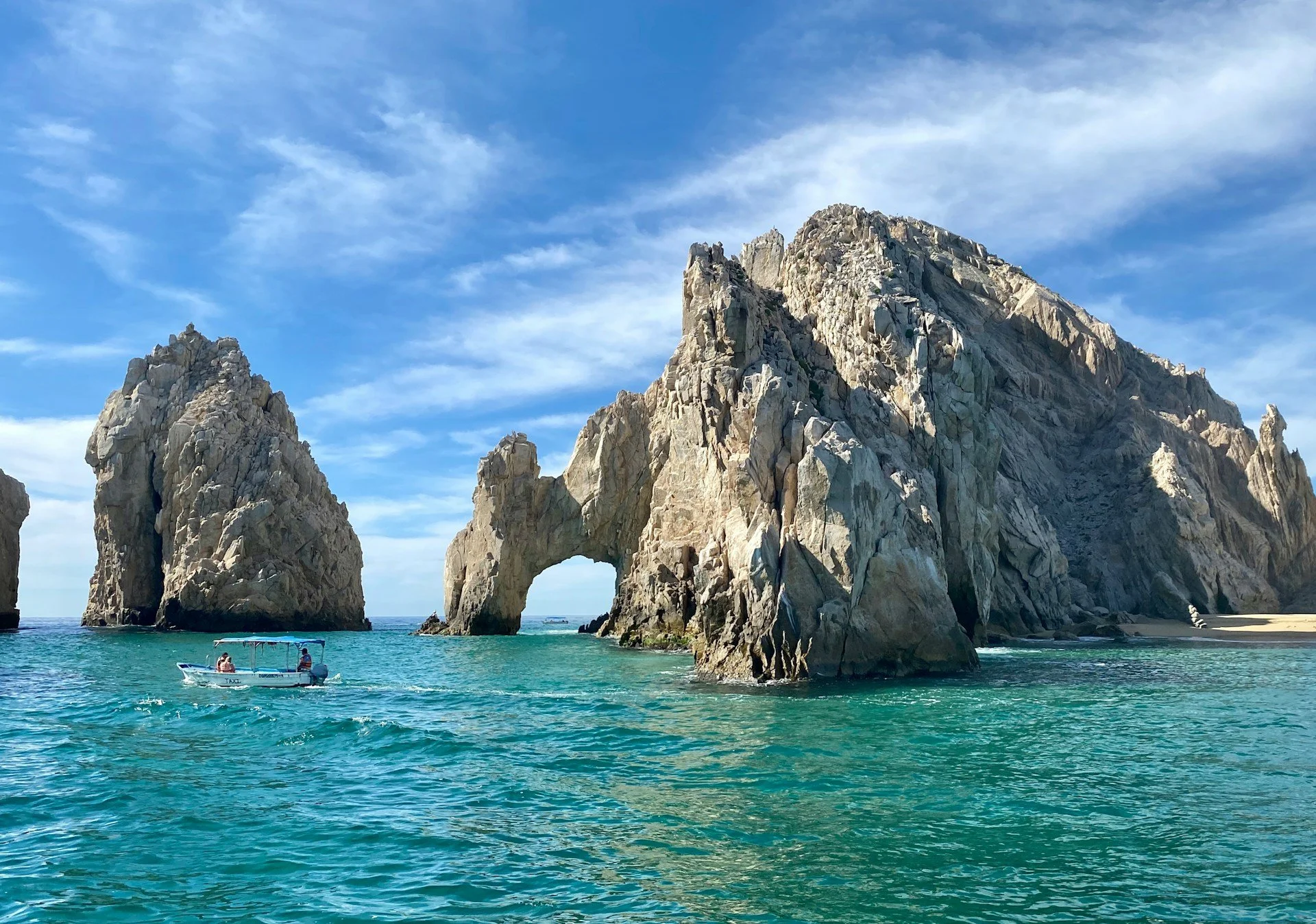 Large rocky coastal formations with an arch and a boat in turquoise water under a partly cloudy sky.