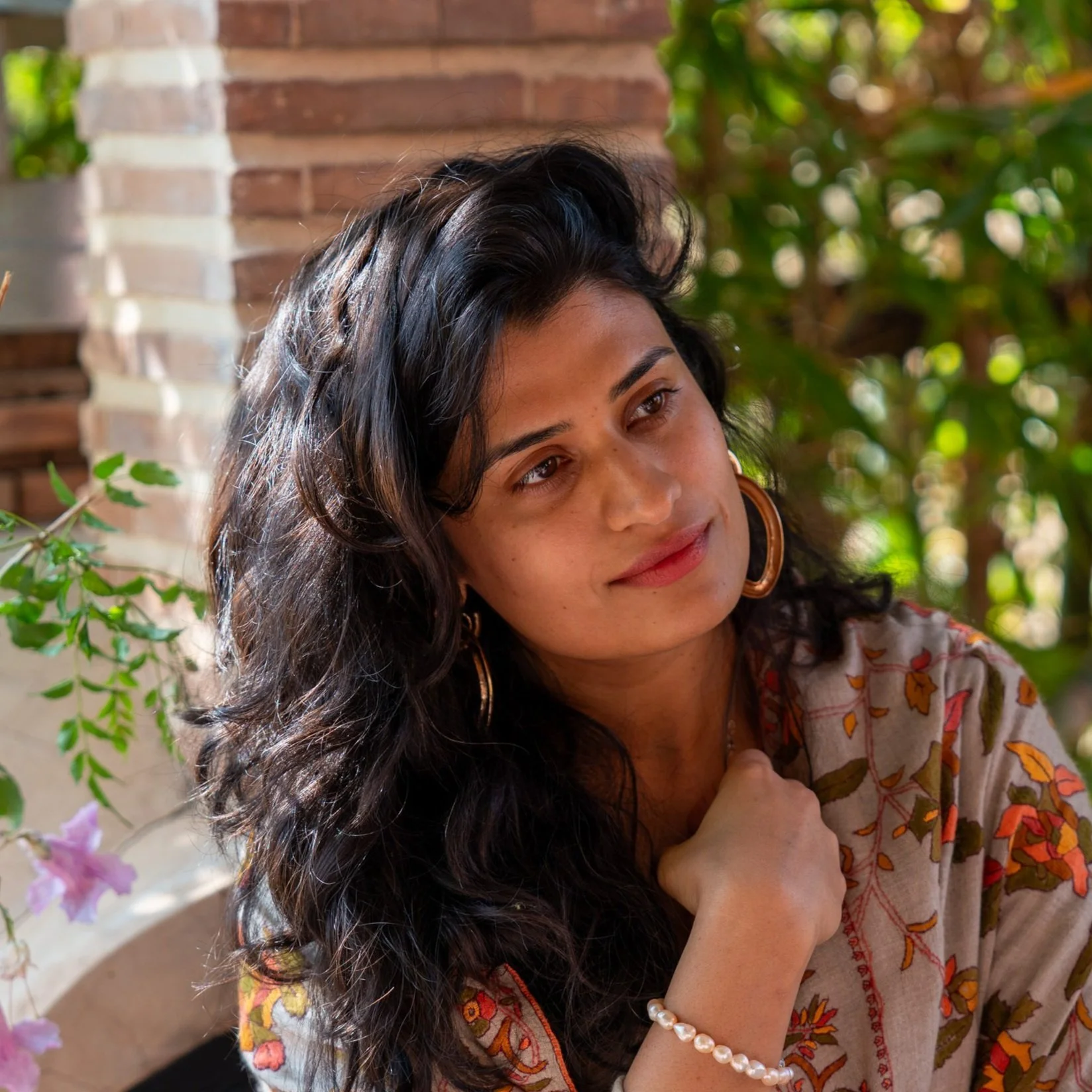 A woman with long black hair, wearing a floral-patterned shawl and gold hoop earrings, sitting in a garden with brick and greenery in the background.