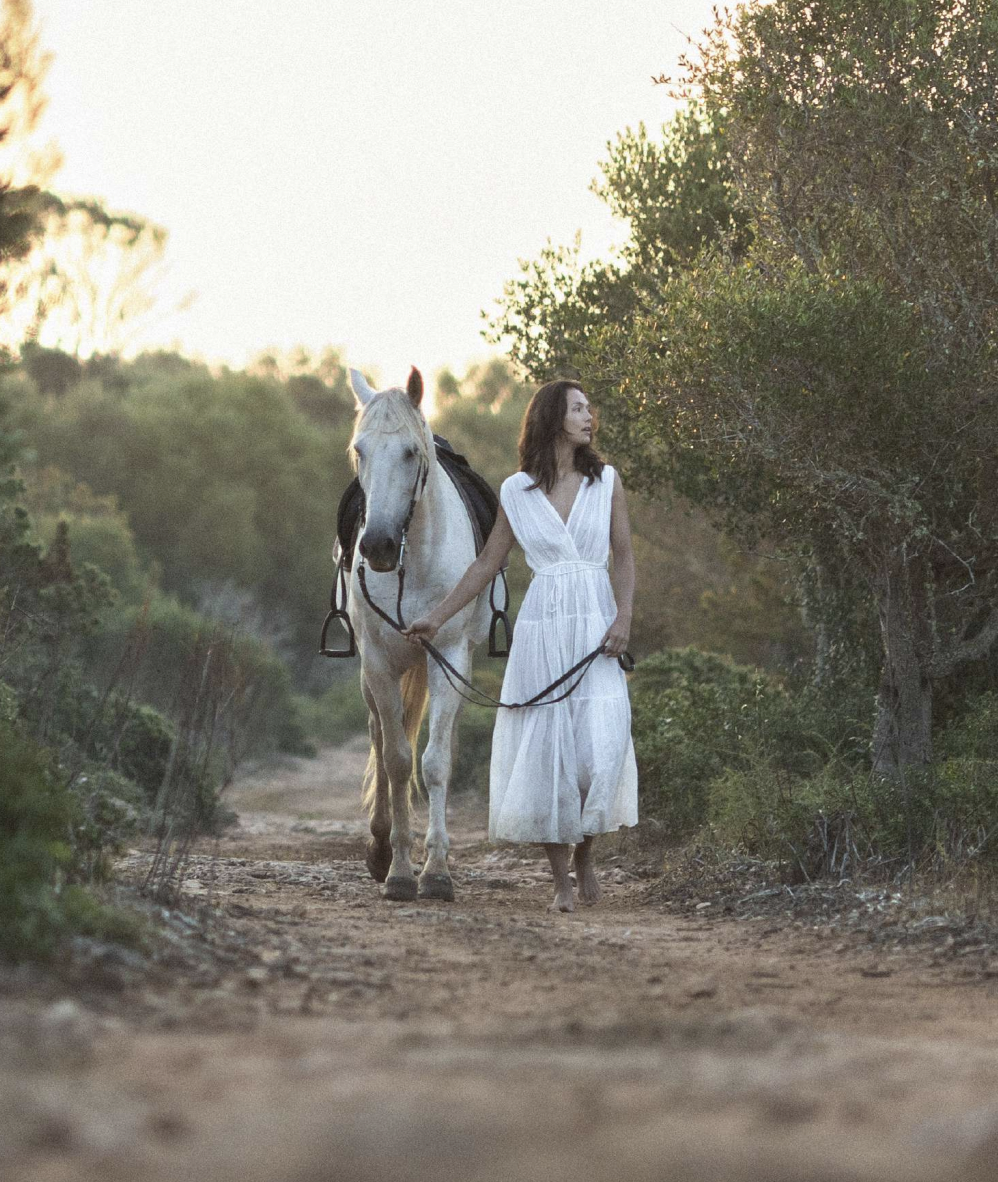 A woman in a white dress walks barefoot on a dirt path next to a white horse, holding its reins, with green trees and bushes around and a soft sunset in the background.