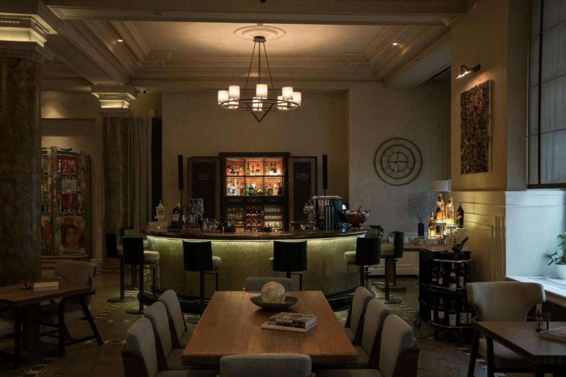 Interior of a stylish bar with a central illuminated curved counter, surrounded by black and white chairs, with shelves of bottles and decorative items behind the bar, warm lighting, and a chandelier hanging from the ceiling.