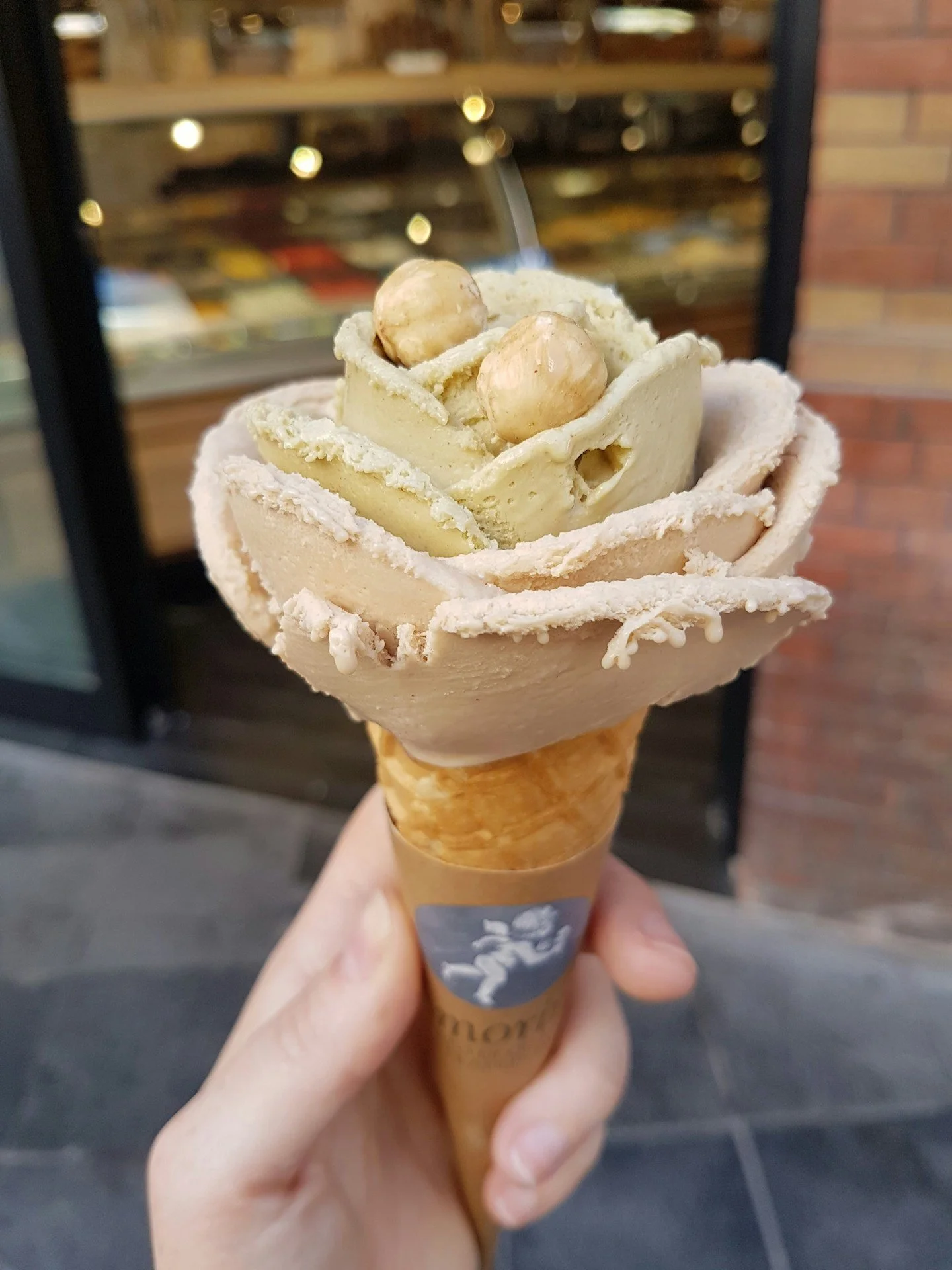Close-up of a gelato cone topped with a swirl of light-colored gelato, hazelnuts, and white chocolate shavings, held outdoors against a blurred background of a brick wall and display case.