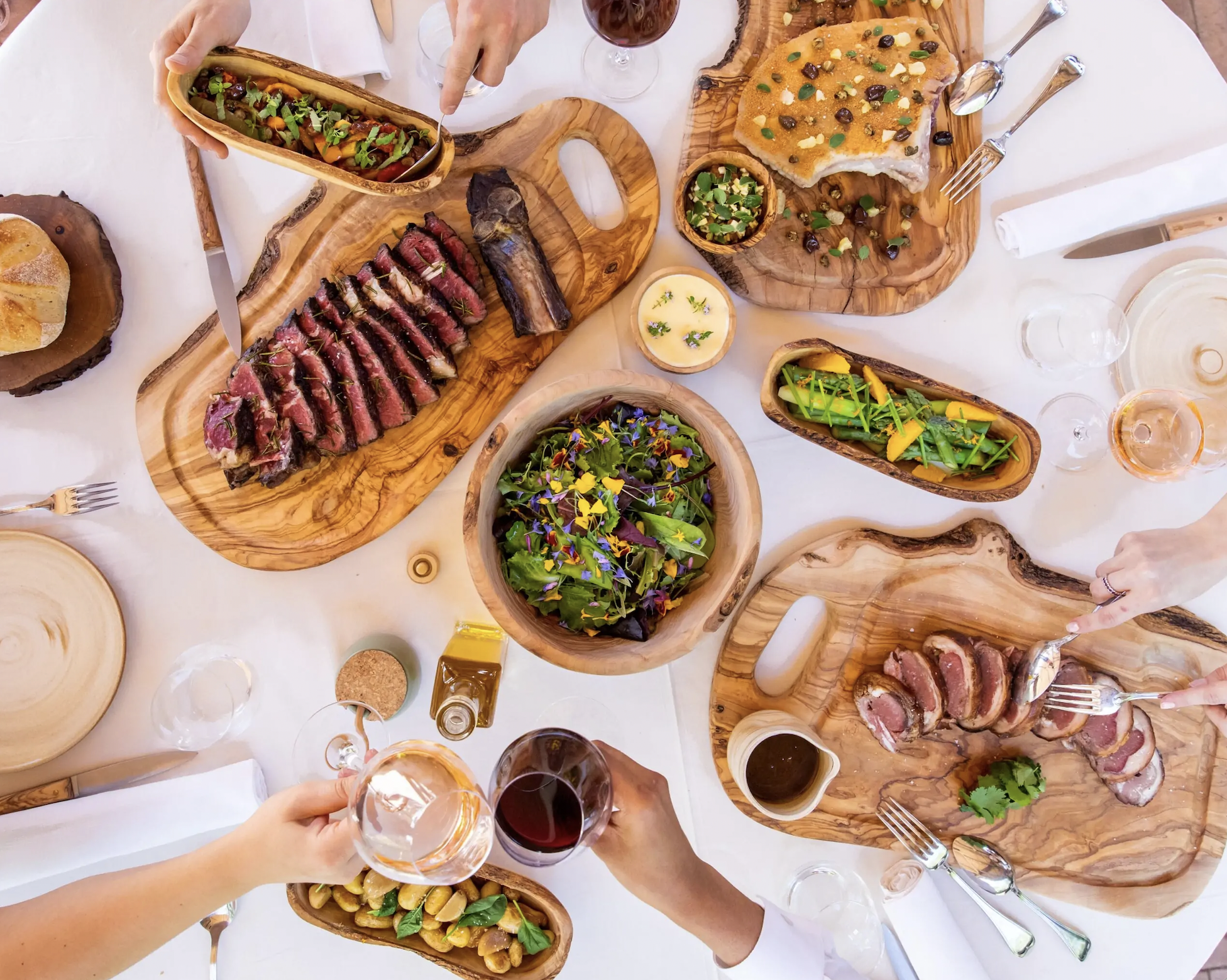Top view of a dining table set with sliced meats, a salad, fresh bread, and wine glasses. The wooden serving boards display vibrant and colorful dishes, surrounded by hands reaching for food and wine.