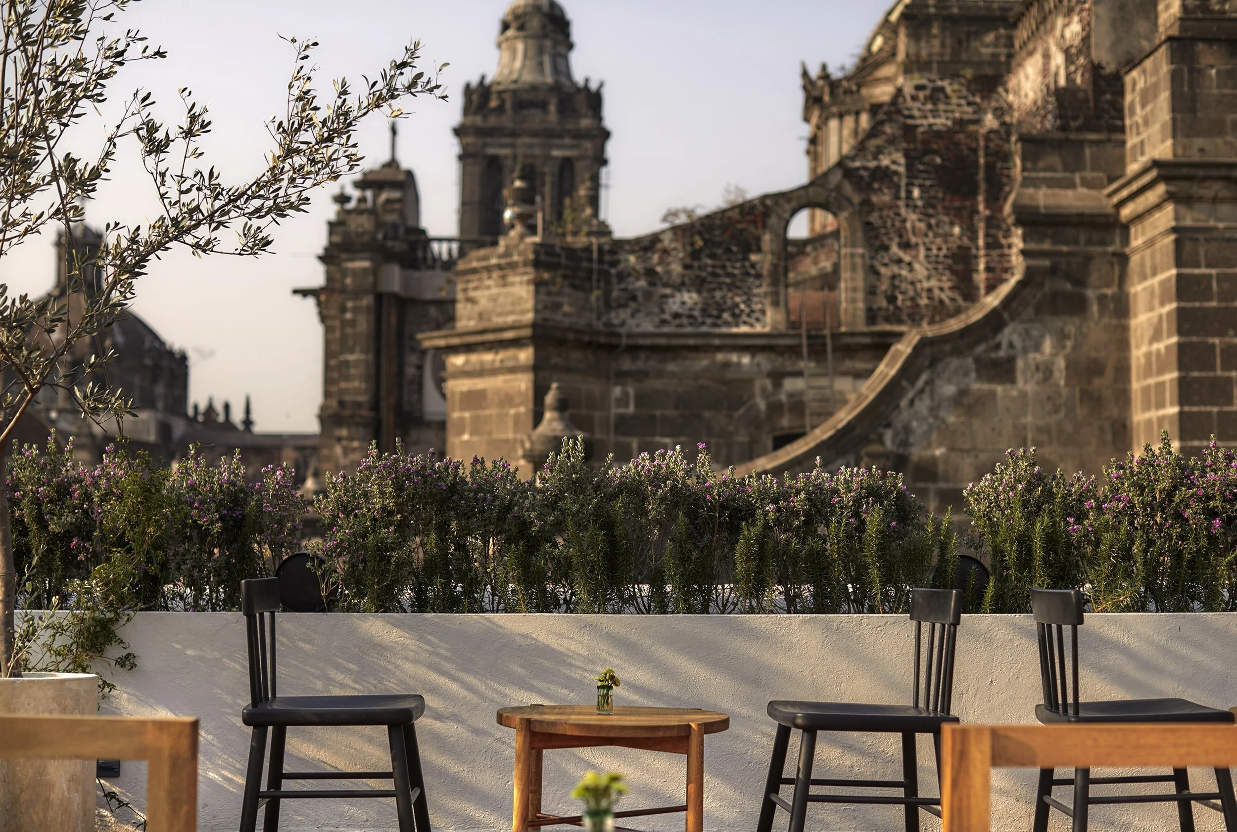Outdoor seating area with three black chairs and a wooden table, surrounded by greenery and purple flowers, with historic stone buildings in the background.