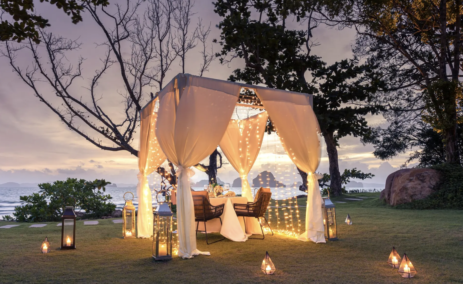 Romantic outdoor dining setup with a canopy and string lights near the ocean at sunset.
