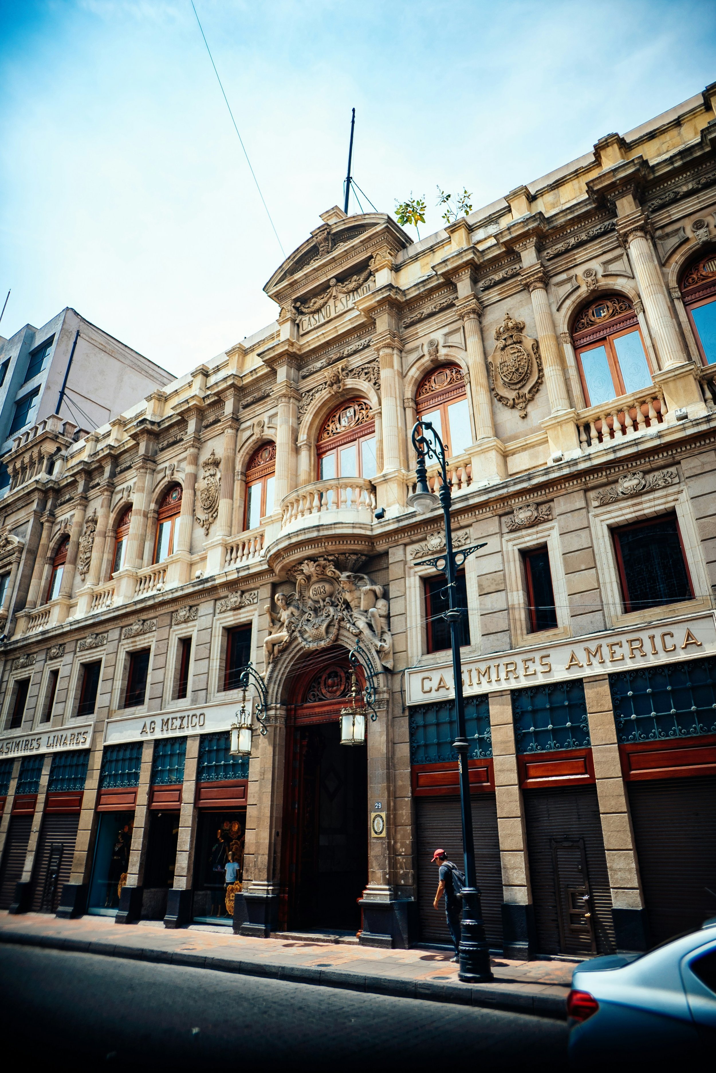 Historic ornate building facade with "Casino Español" inscription, featuring columns, decorative carvings, and arched windows, alongside street lamps and a pedestrian on a sunny day.