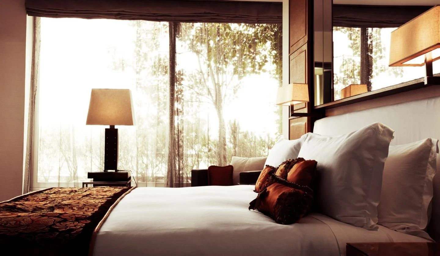 Elegant bedroom with white bedding, decorative pillows, large windows with sheer curtains, and warm lighting.