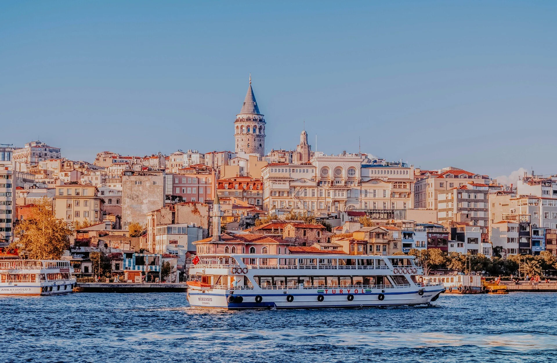 A view of a city skyline with various buildings and the iconic Galata Tower in the background, with a boat in the water in the foreground.