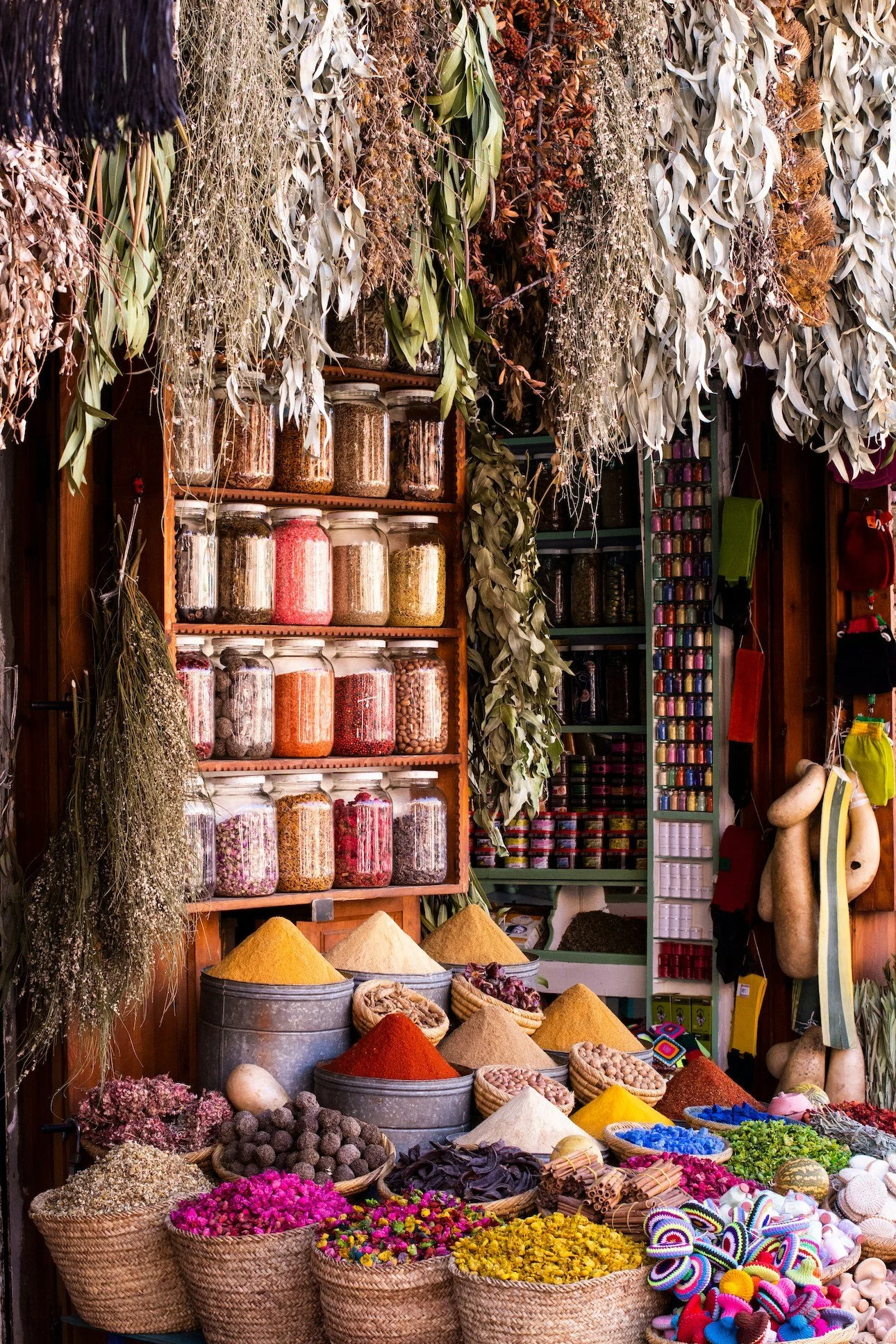 A colorful market stall with jars of spices and dried herbs on shelves, and baskets of vibrant dried flowers, powders, and craft materials on the table.