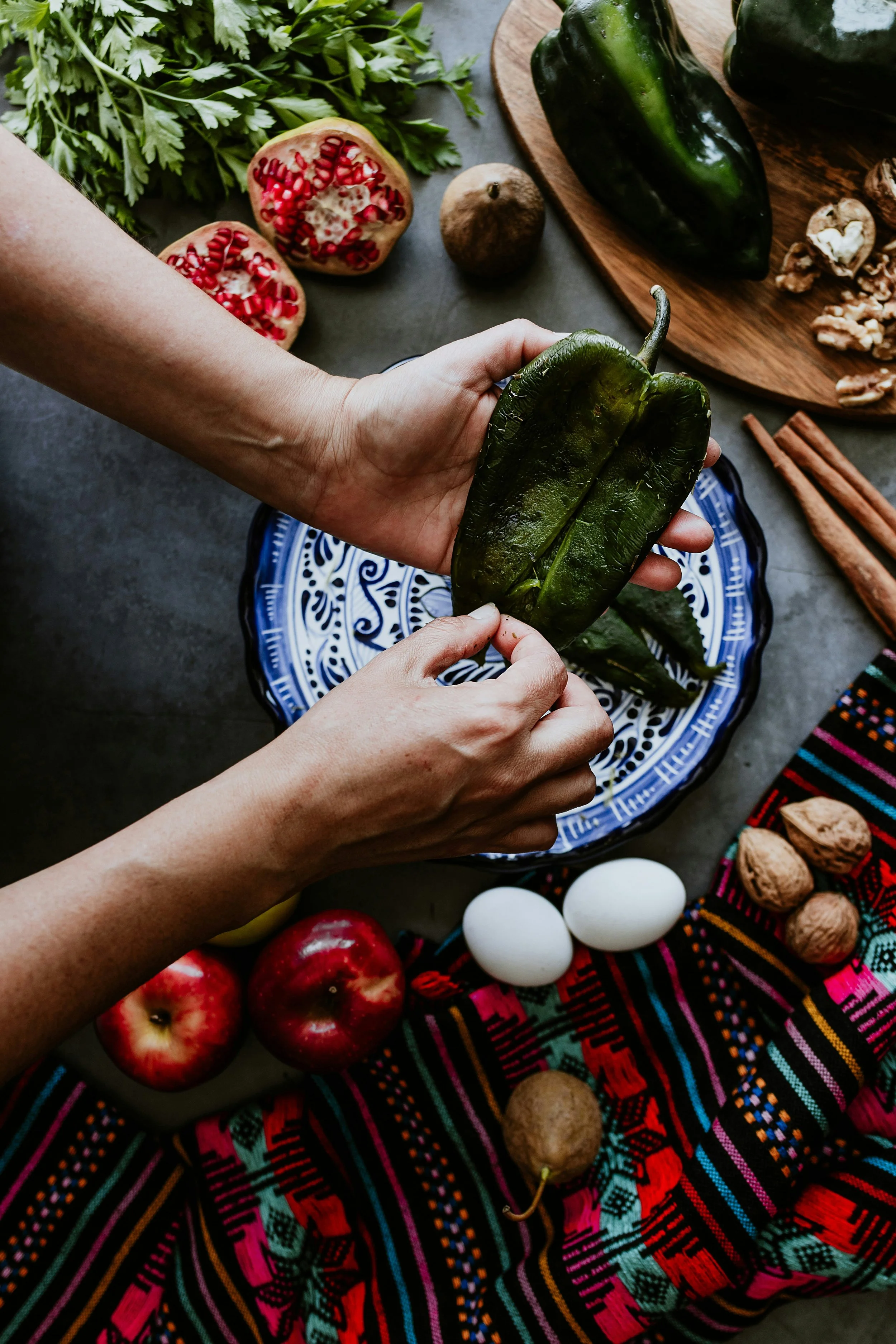 Person holding roasted poblano pepper over a blue patterned plate, surrounded by ingredients like pomegranates, walnuts, eggs, apples, leafy greens, and a colorful textile.